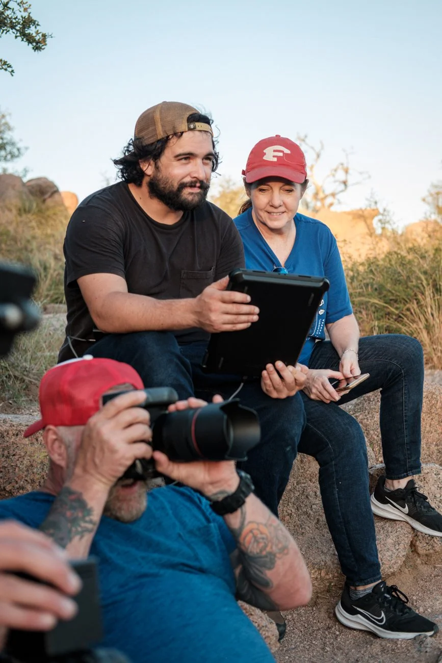 Three people outdoors, sitting on rocks, looking at a tablet and smiling. One person has a camera, another is holding a smartphone. They seem to be filming or photographing something during sunset or sunrise.