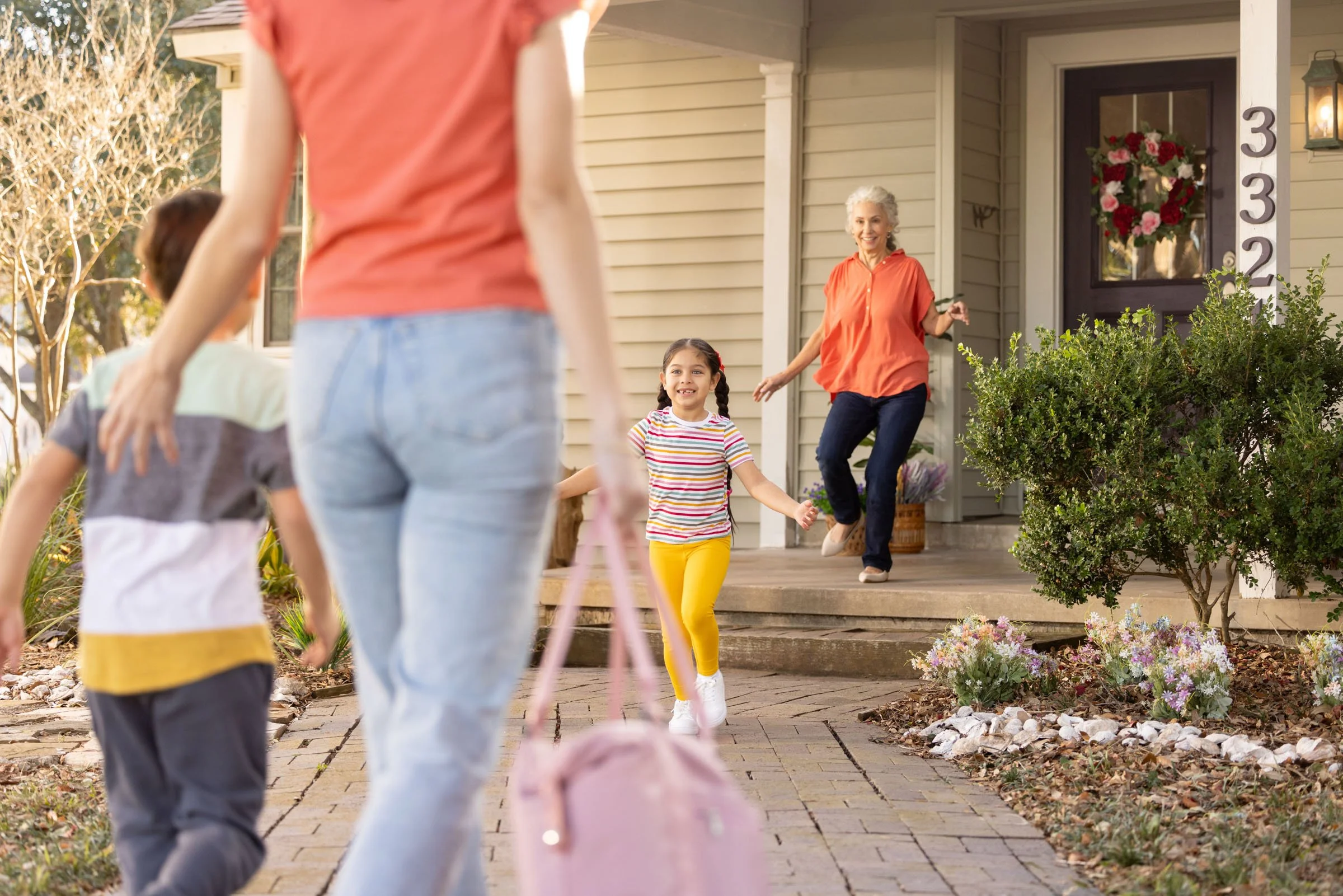 A woman and two children walking towards a smiling elderly woman on the front porch of a house decorated with a wreath. The scene is outdoors during daytime.