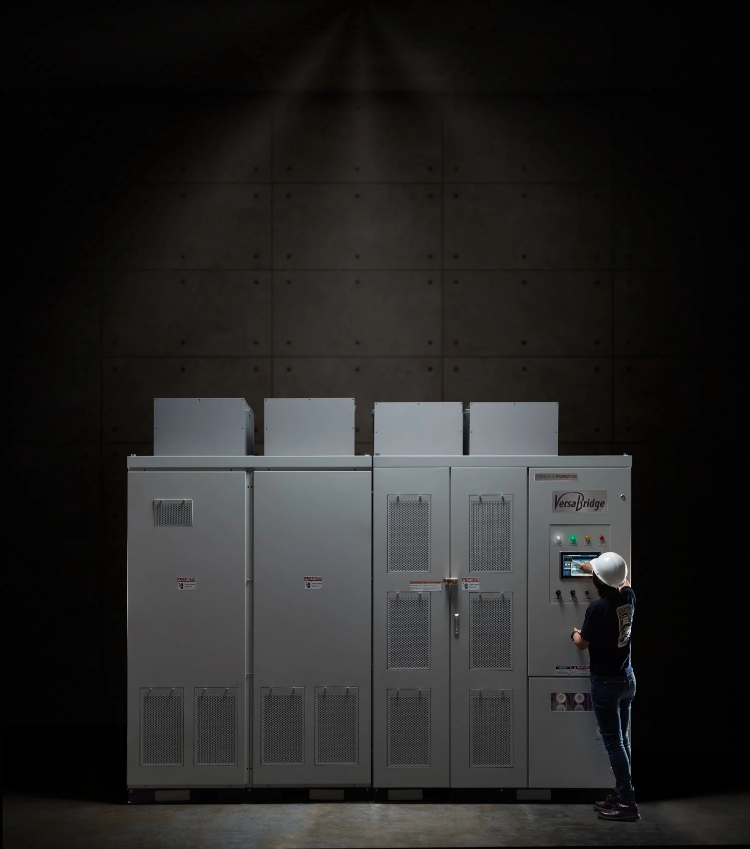 A person wearing a white safety helmet is operating a control panel on a large electrical cabinet labeled 'VersaBridge' in a dark room with a textured wall in the background.