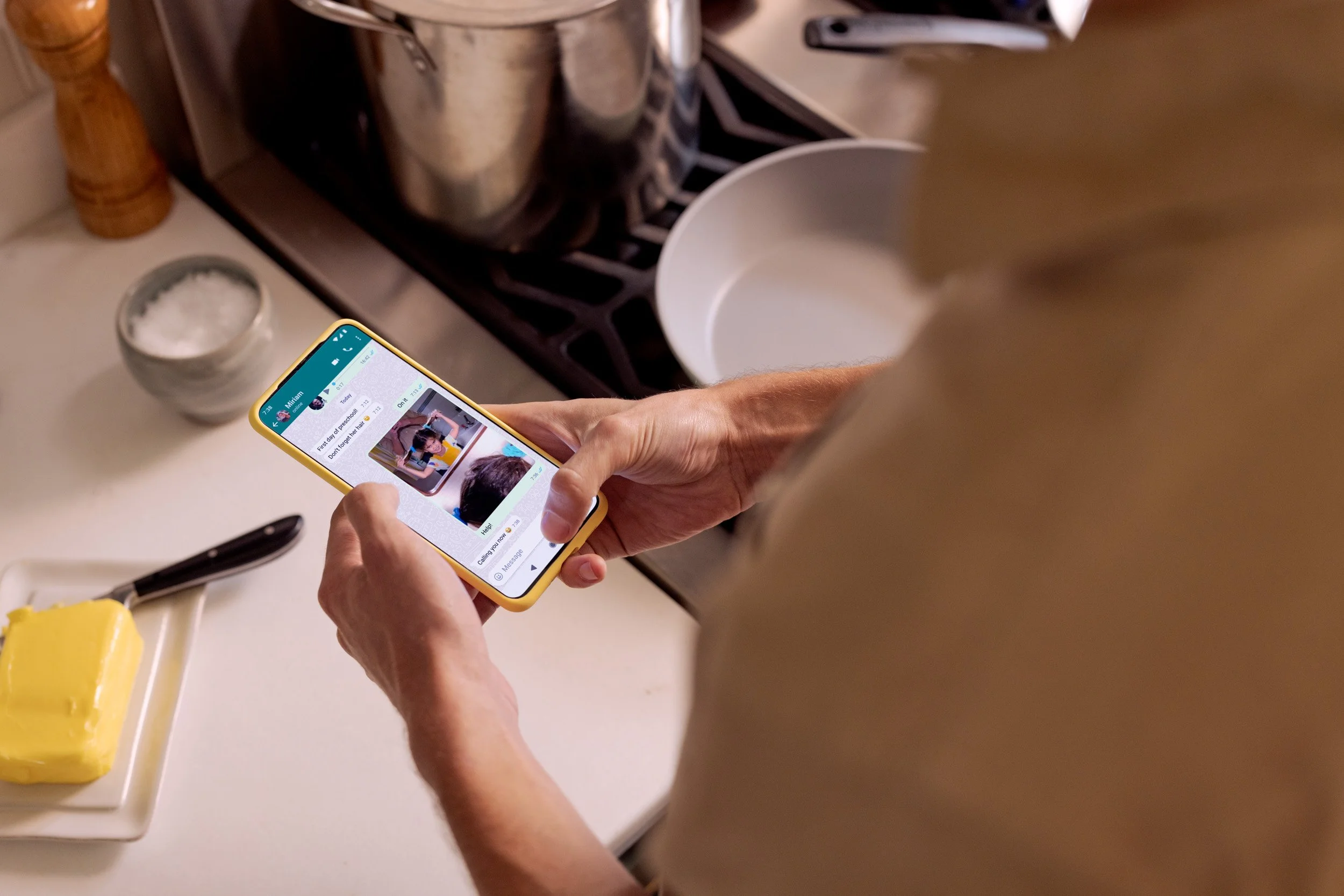 Person holding a smartphone above a kitchen countertop, showing a WhatsApp conversation with a picture of a young girl. The countertop has a white dish, a yellow block of butter, a black-handled knife, a white tray with some yellow substance, a glass