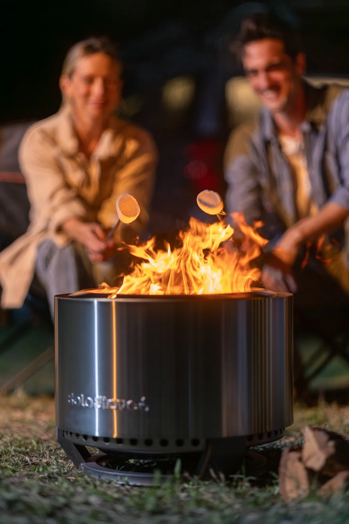 Two people sitting outdoors at night, roasting marshmallows over a fire in a portable fire pit.