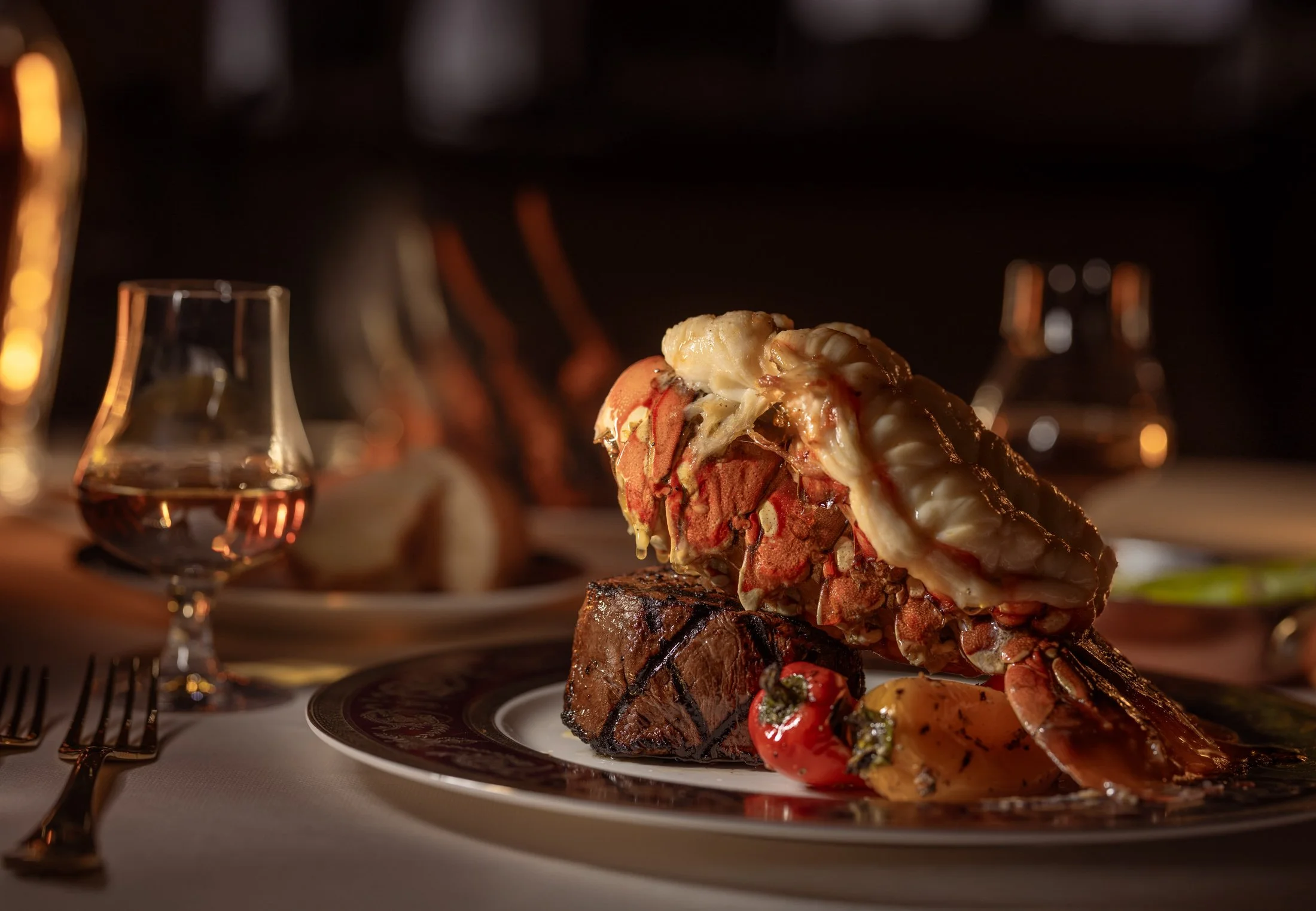 A gourmet meal featuring a large lobster tail, grilled steak, cherry tomatoes, and roasted potatoes on an elegant plate with glassware and utensils on the table in a dimly lit restaurant setting.