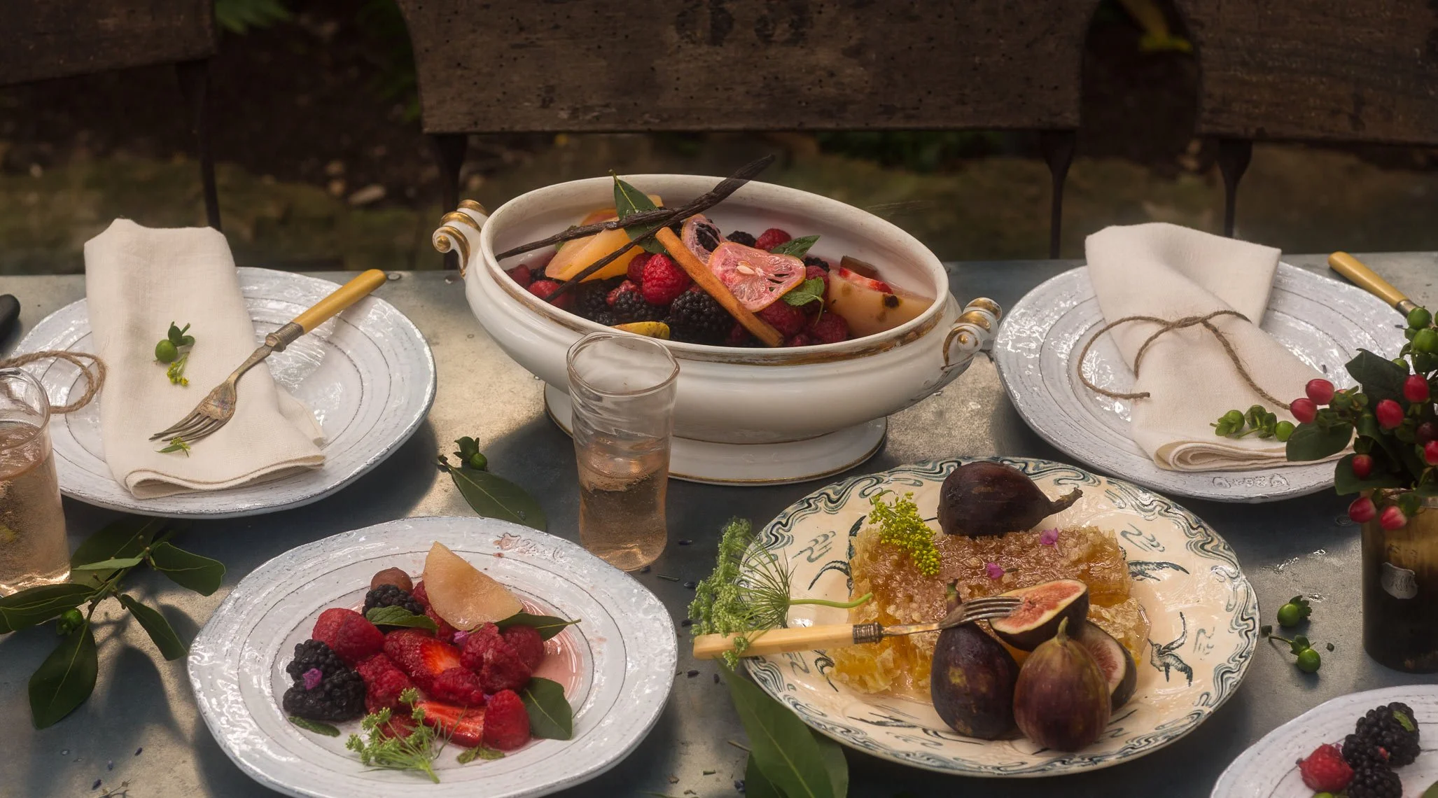 A table set for a meal with various plates of fresh berries, figs, and desserts, with cloth napkins, forks, and glasses of water, outdoors with greenery in the background.