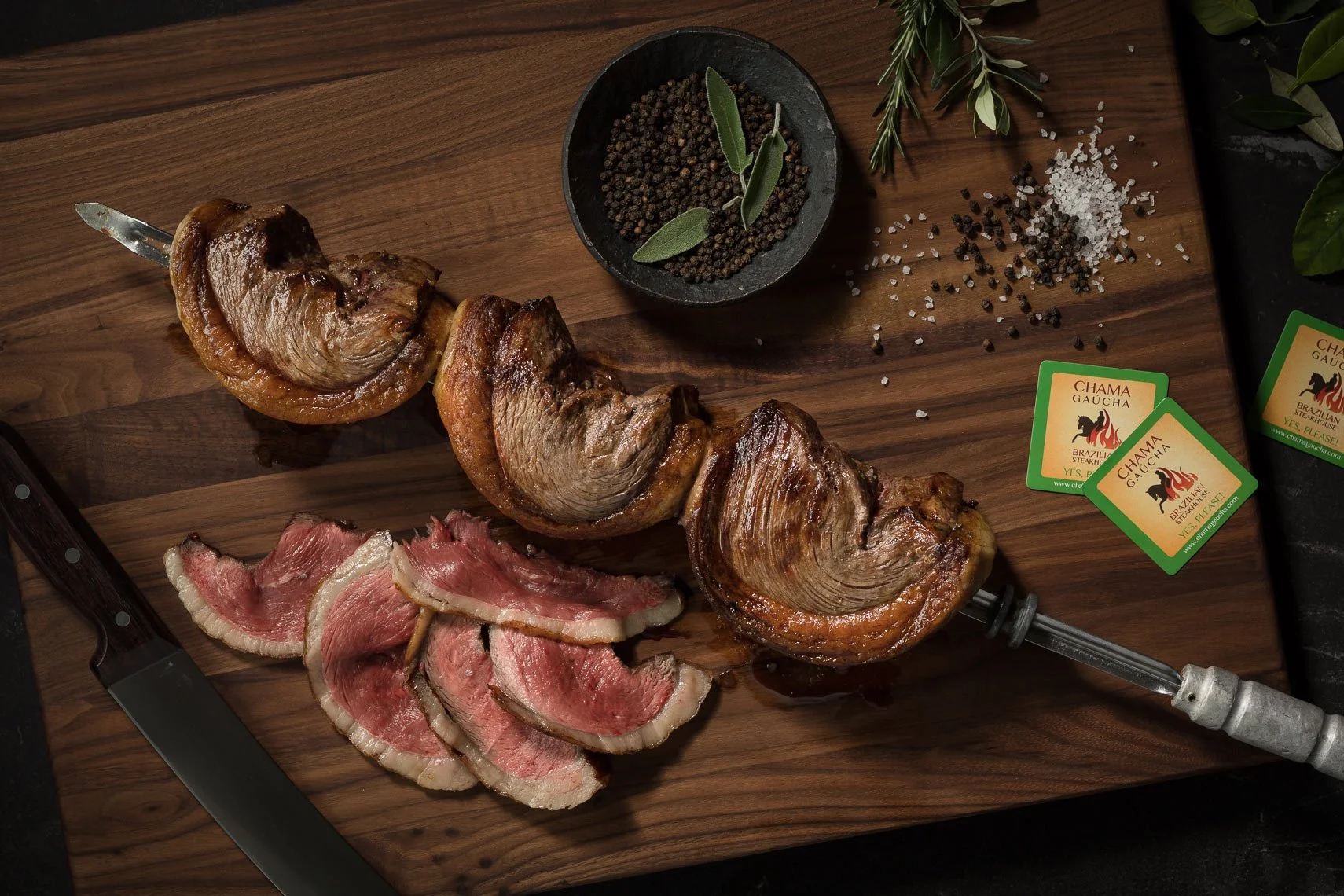 Sliced raw beef with bacon, grilled beef heart or similar organ meat, seasoned salt, black pepper, and fresh herbs on a wooden cutting board, with a knife, a meat injector, and Chama Gaucha steakhouse tags nearby.