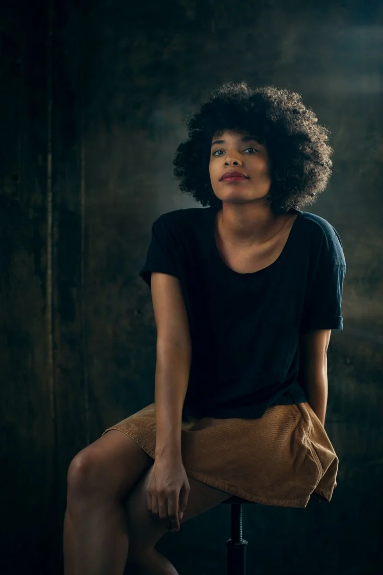 A woman with curly hair wearing a black t-shirt and brown shorts sitting on a stool against a dark background.