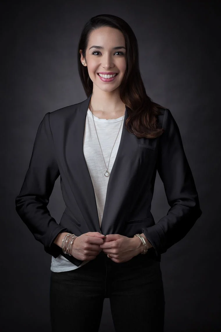 A woman with long dark hair, smiling and wearing a black blazer over a white t-shirt, standing against a dark background.