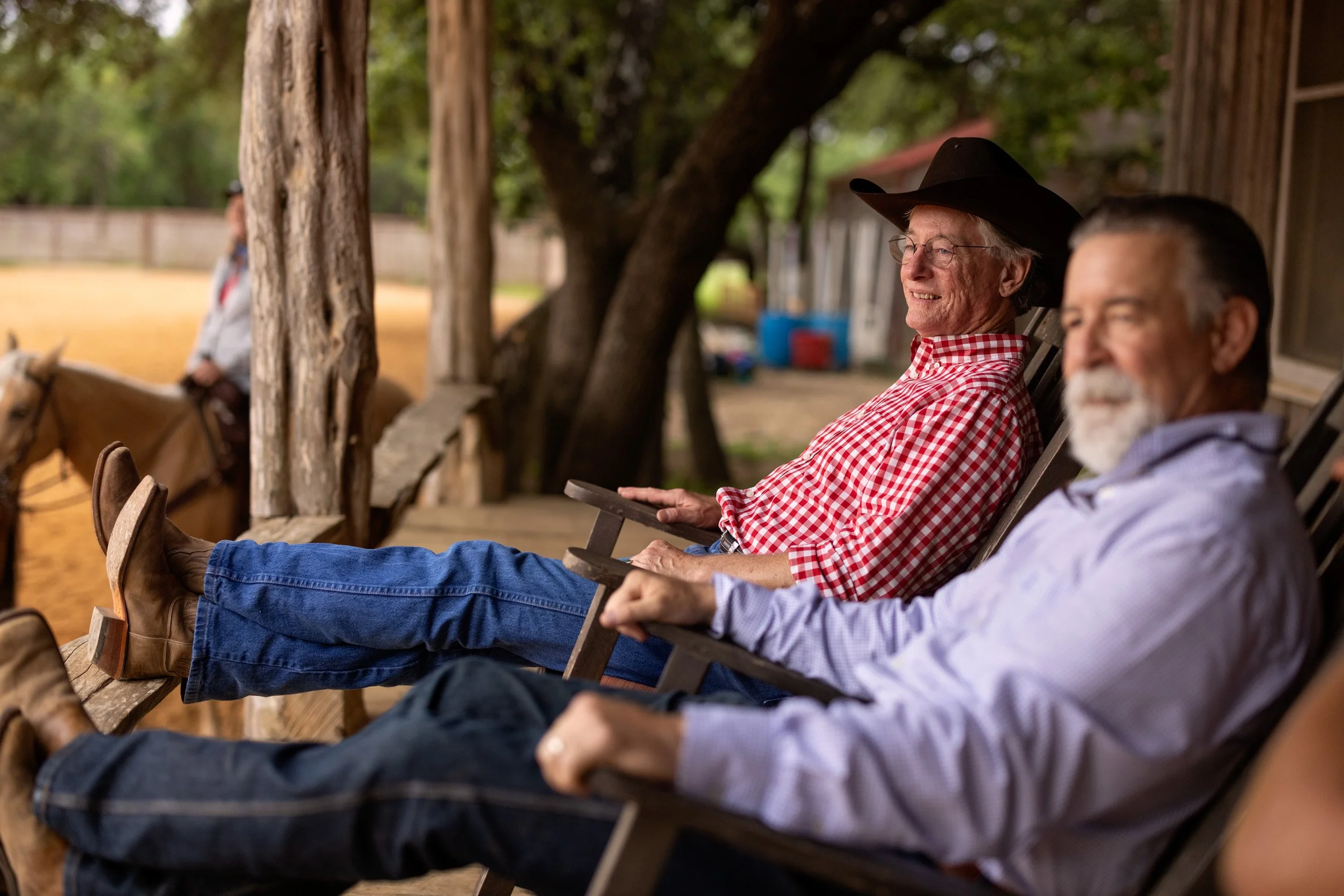 Two elderly men sitting in lawn chairs outdoors under trees, smiling, one wearing a cowboy hat and glasses, the other with gray hair and a beard, both dressed casually.