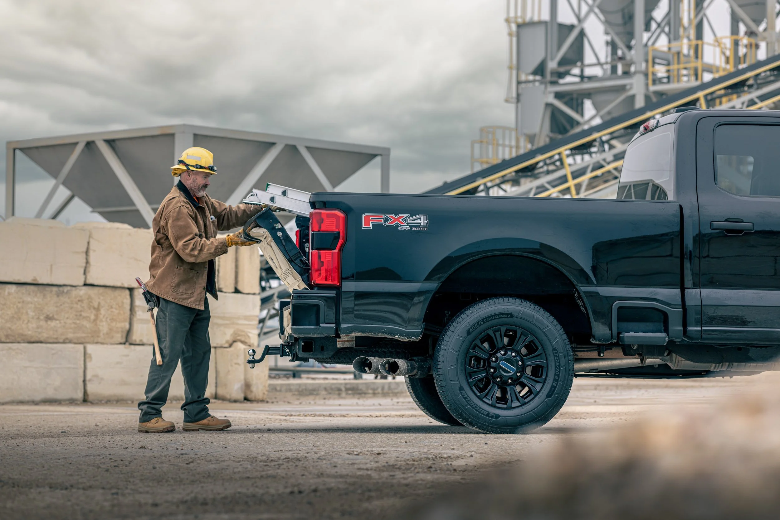 A construction worker in a yellow hard hat and brown jacket loading a metal object into the back of a black pickup truck with an FX4 logo on the tailgate. The background features construction equipment and a cloudy sky.