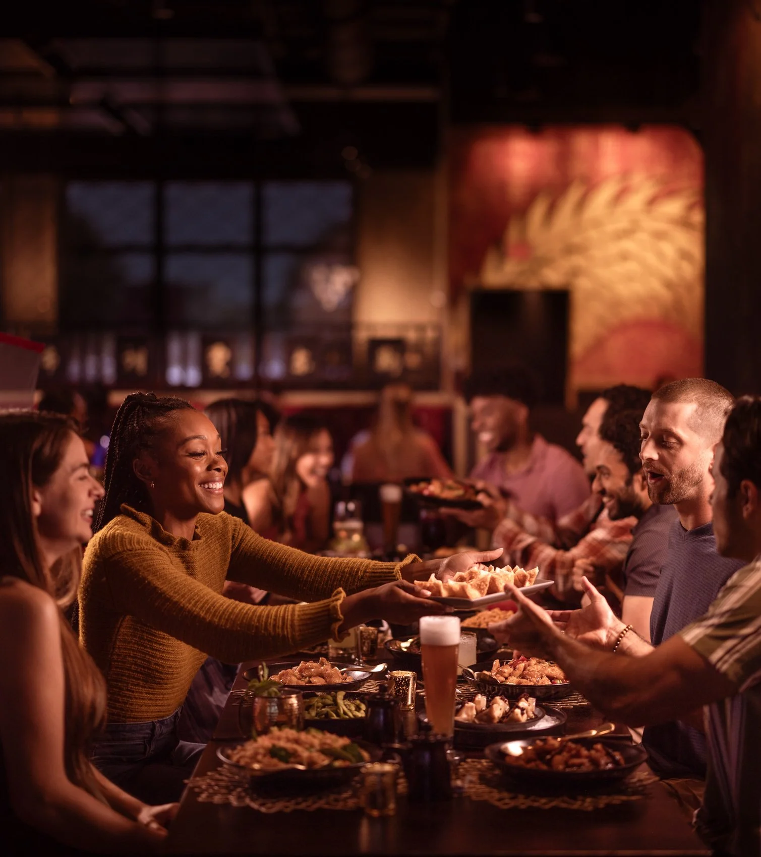 People enjoying a dinner in a restaurant, with a woman passing food to a man across the table, and other diners smiling and chatting in the background.