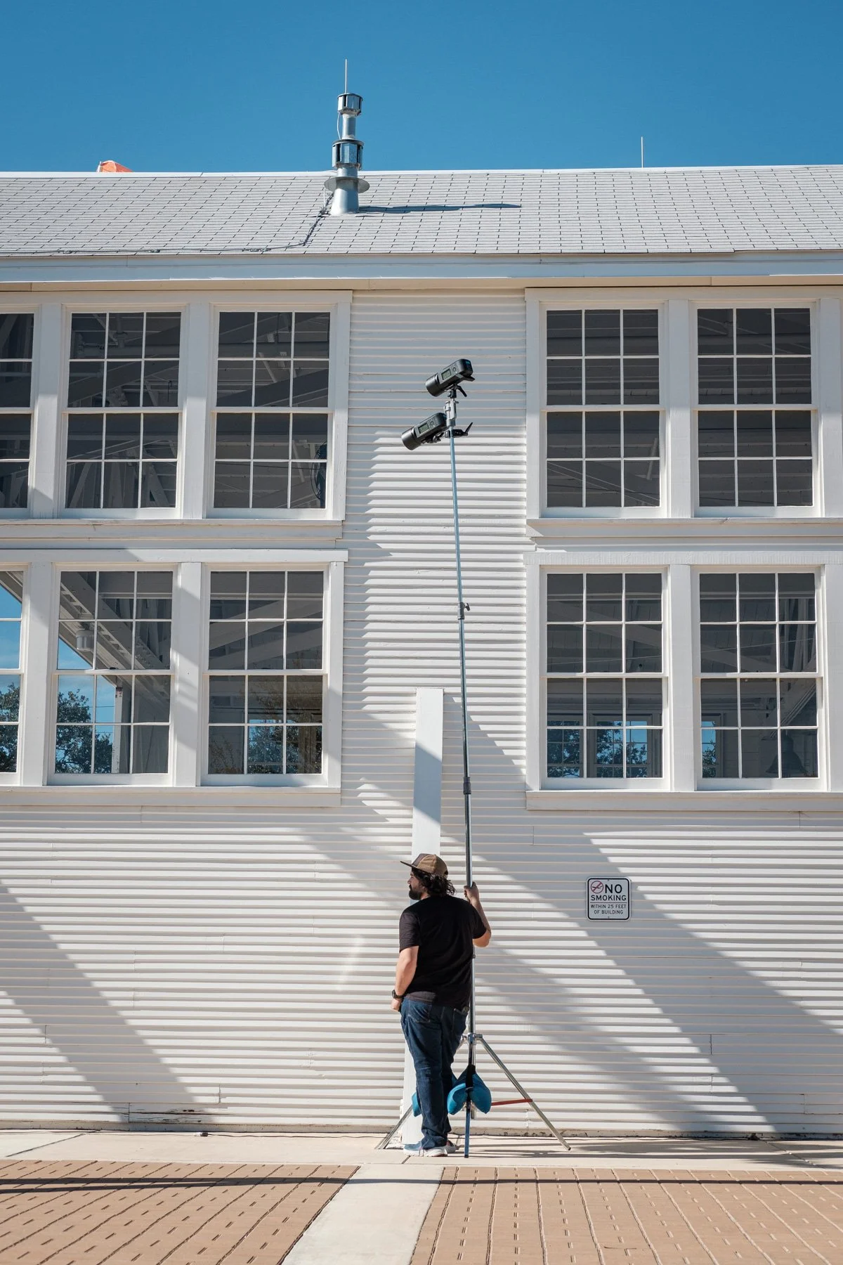 A man standing outside a white building with large windows, holding a pole with two cameras attached, filming a scene.