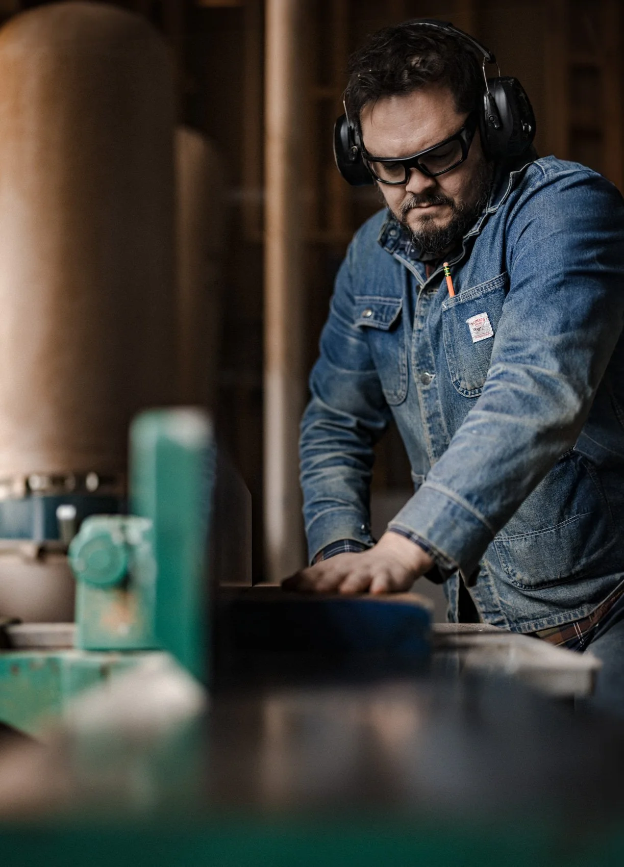 A man wearing glasses, headphones, and a denim jacket working on a woodworking project in a workshop.
