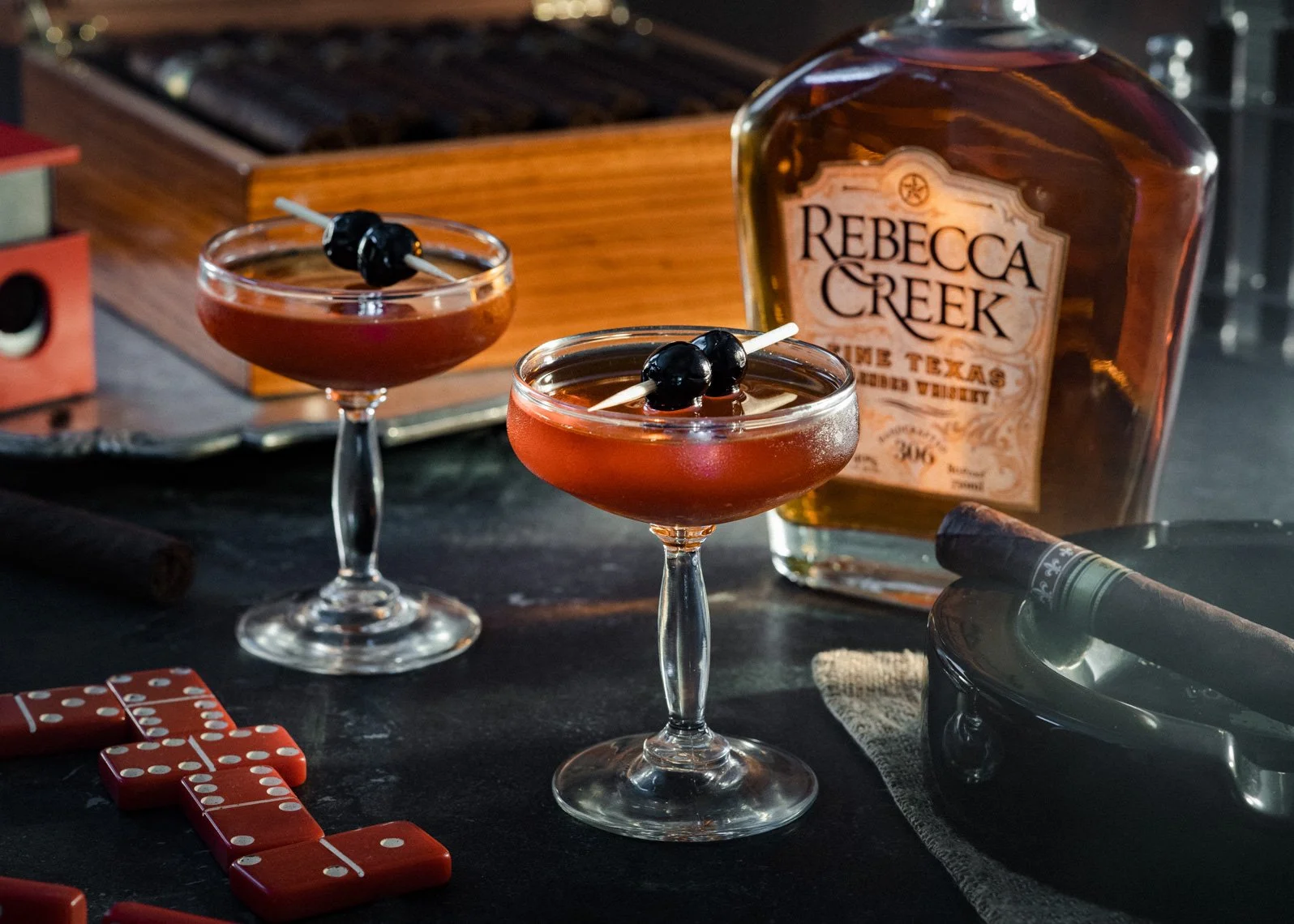 Two cocktails with dark cherry garnishes, a bottle of Rebecca Creek bourbon, dominoes, a cigar, and a cash register on a dark table.