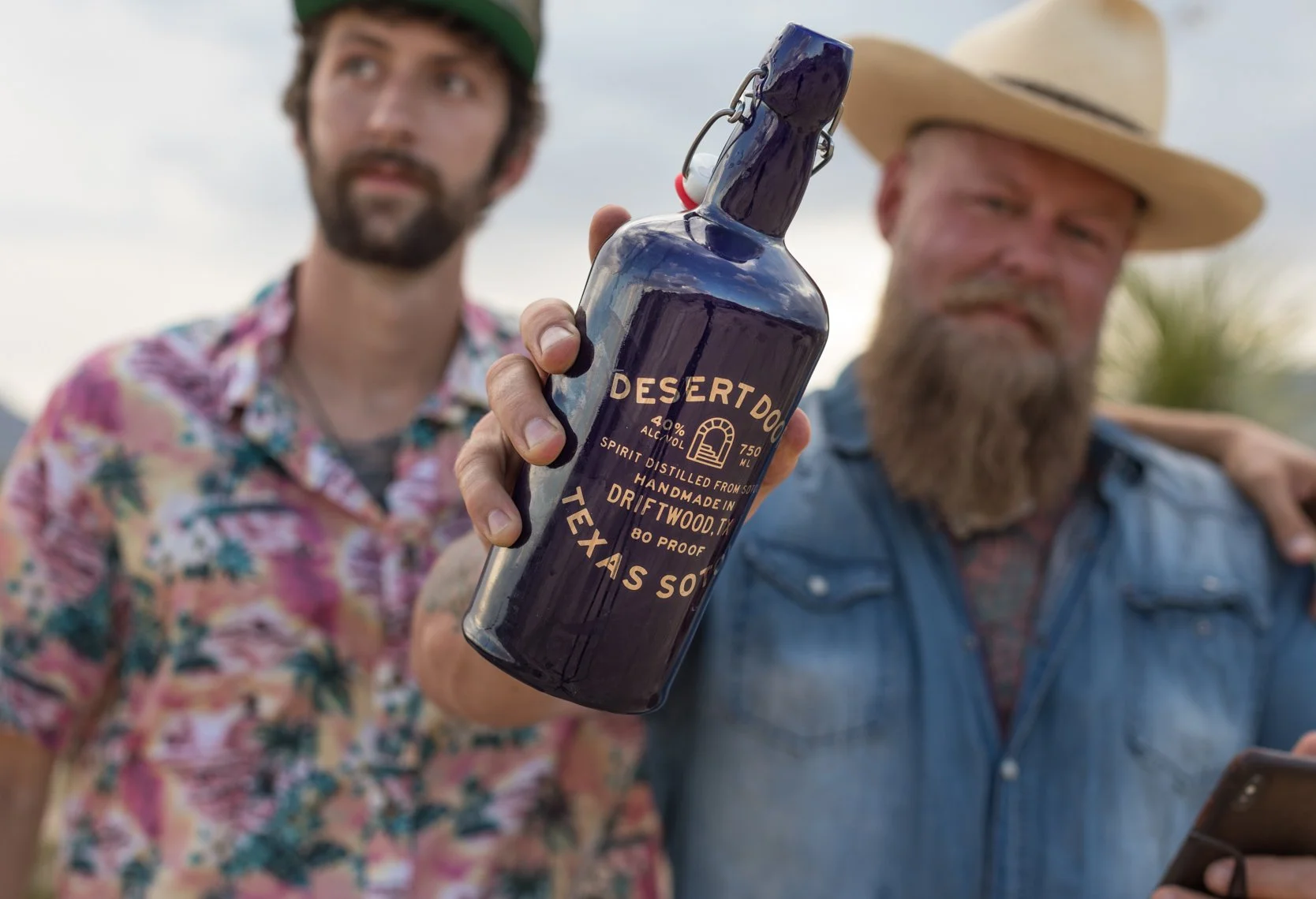 Two men outdoors, one wearing a floral shirt and a green cap, the other with a beard wearing a straw hat and denim jacket, holding a blue bottle of Desert Door Texas Sotol spirit toward the camera, with one man placing his arm around the other's shou
