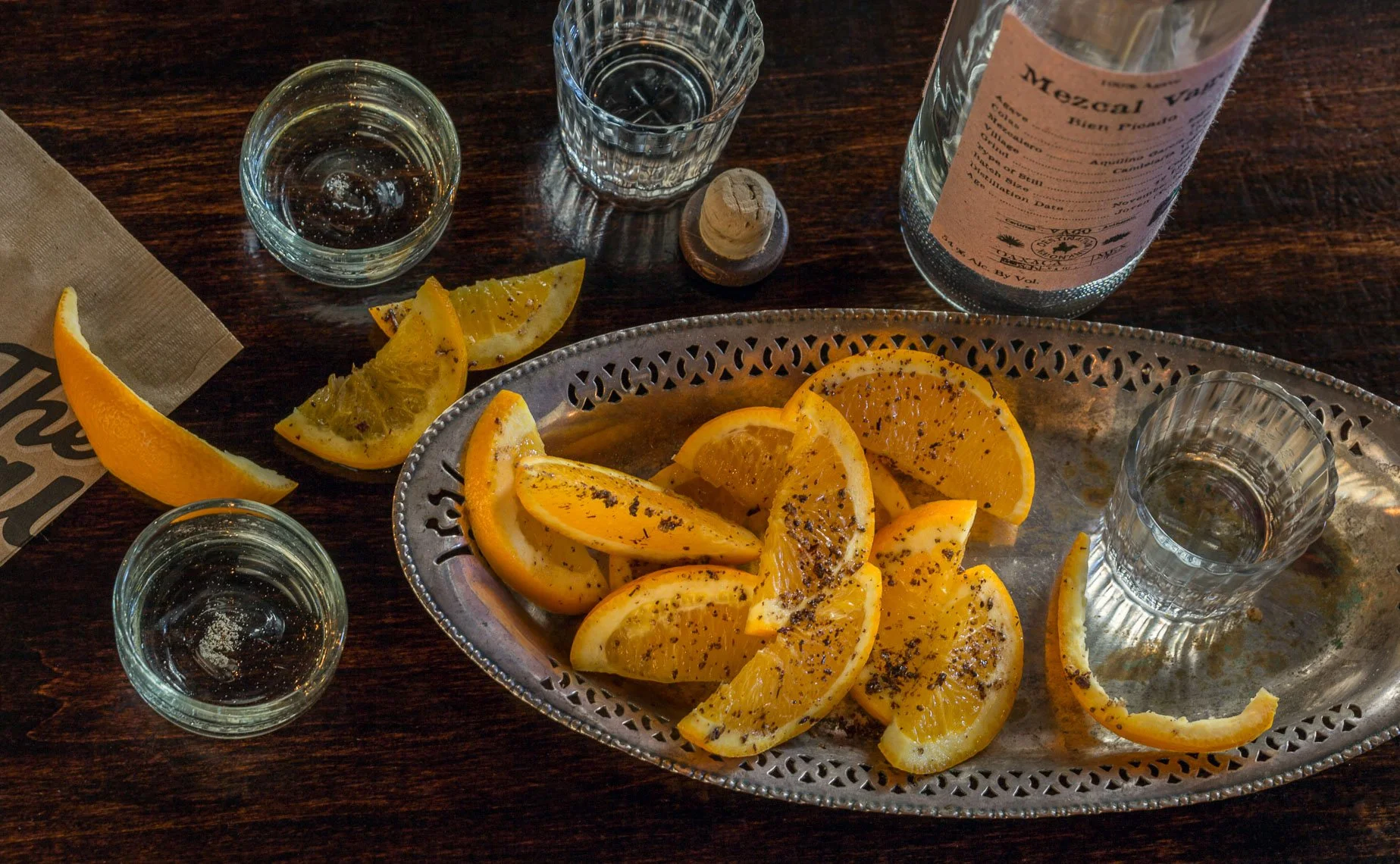 Sliced oranges on a silver tray, some sprinkled with black pepper, alongside lemon wedges, on a dark wooden table with glasses of water and a wine bottle.