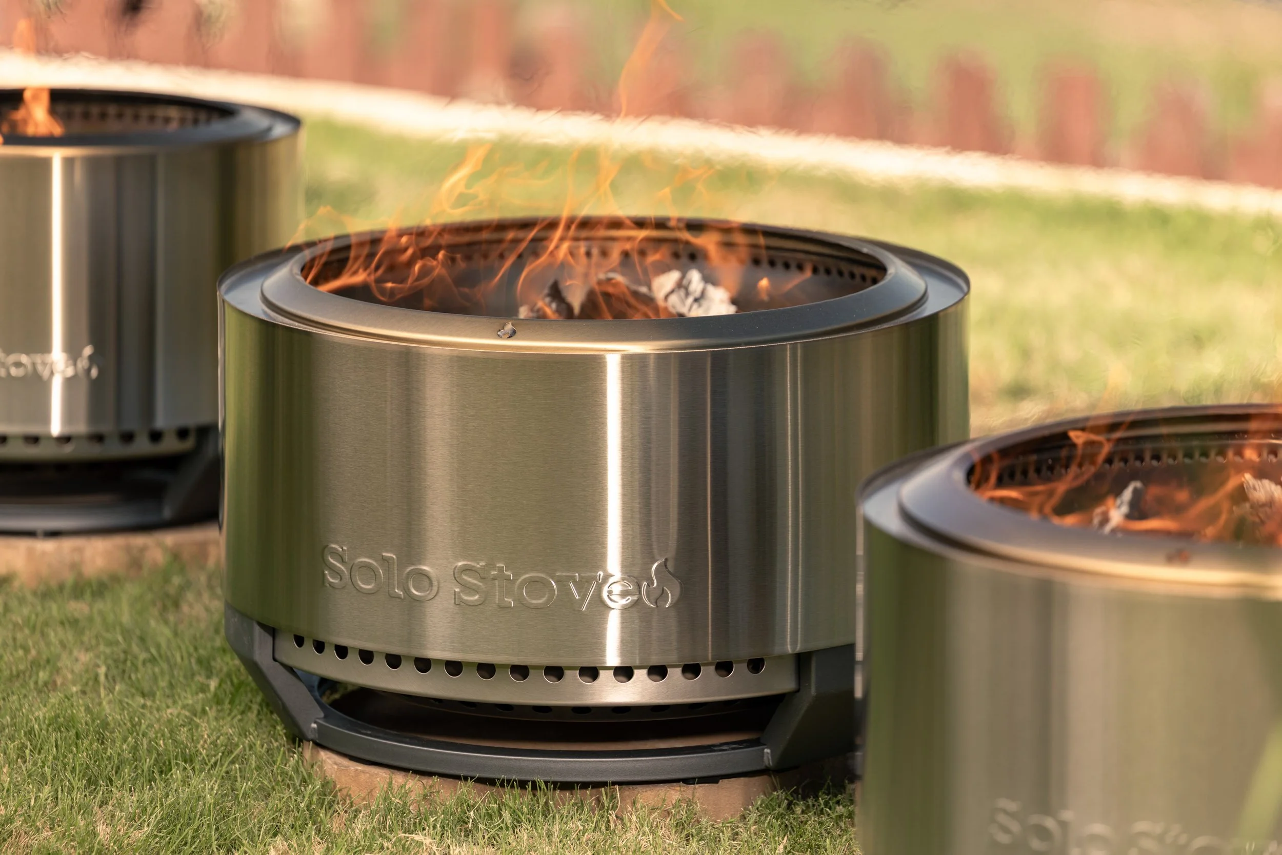 Stainless steel solo stove outdoor fire pit with flames and wood inside, on a grassy surface, with a blurred outdoor background.