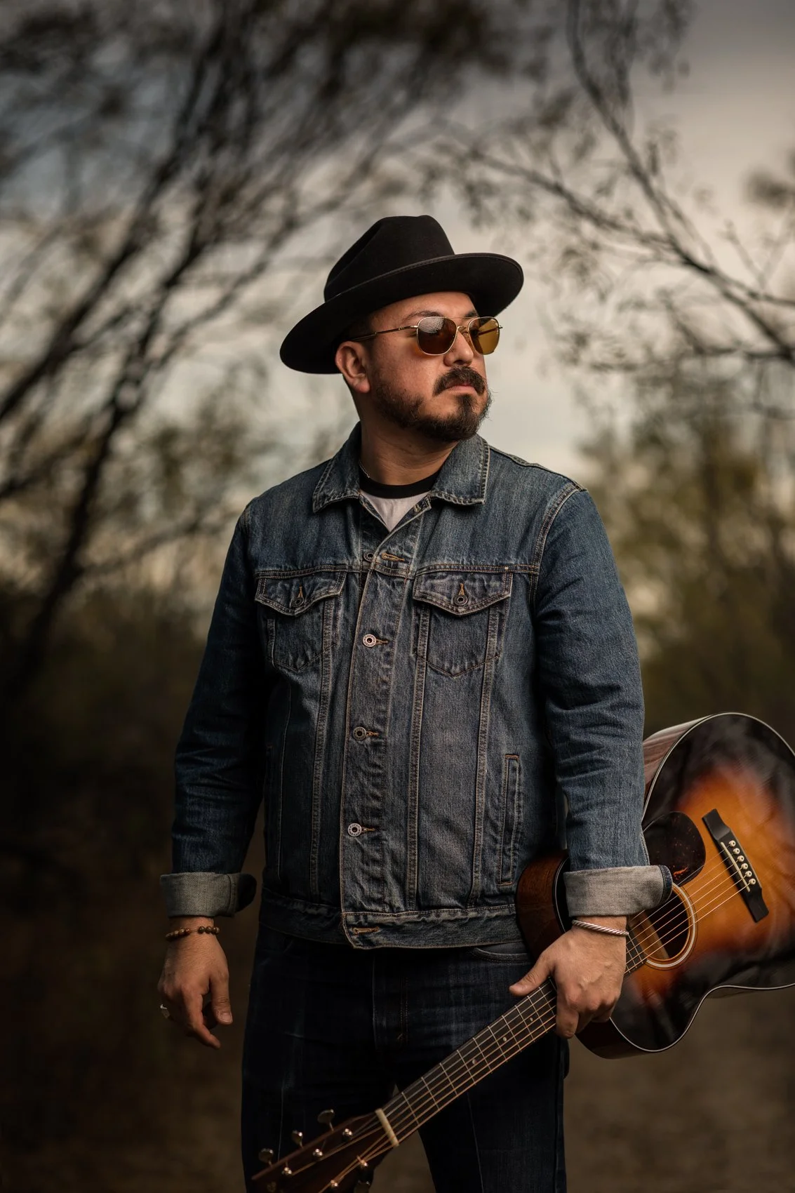 A man with a beard and sunglasses holding an acoustic guitar outdoors, wearing a denim jacket and a black hat, with trees and a cloudy sky in the background.