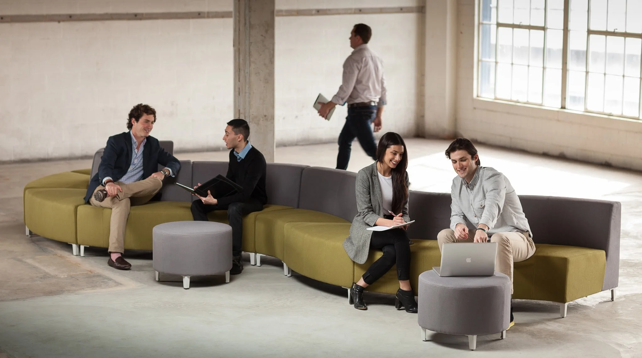 Five young adults in business casual attire are sitting and chatting on a modern, curved sofa in a spacious, industrial-style lounge with large windows.