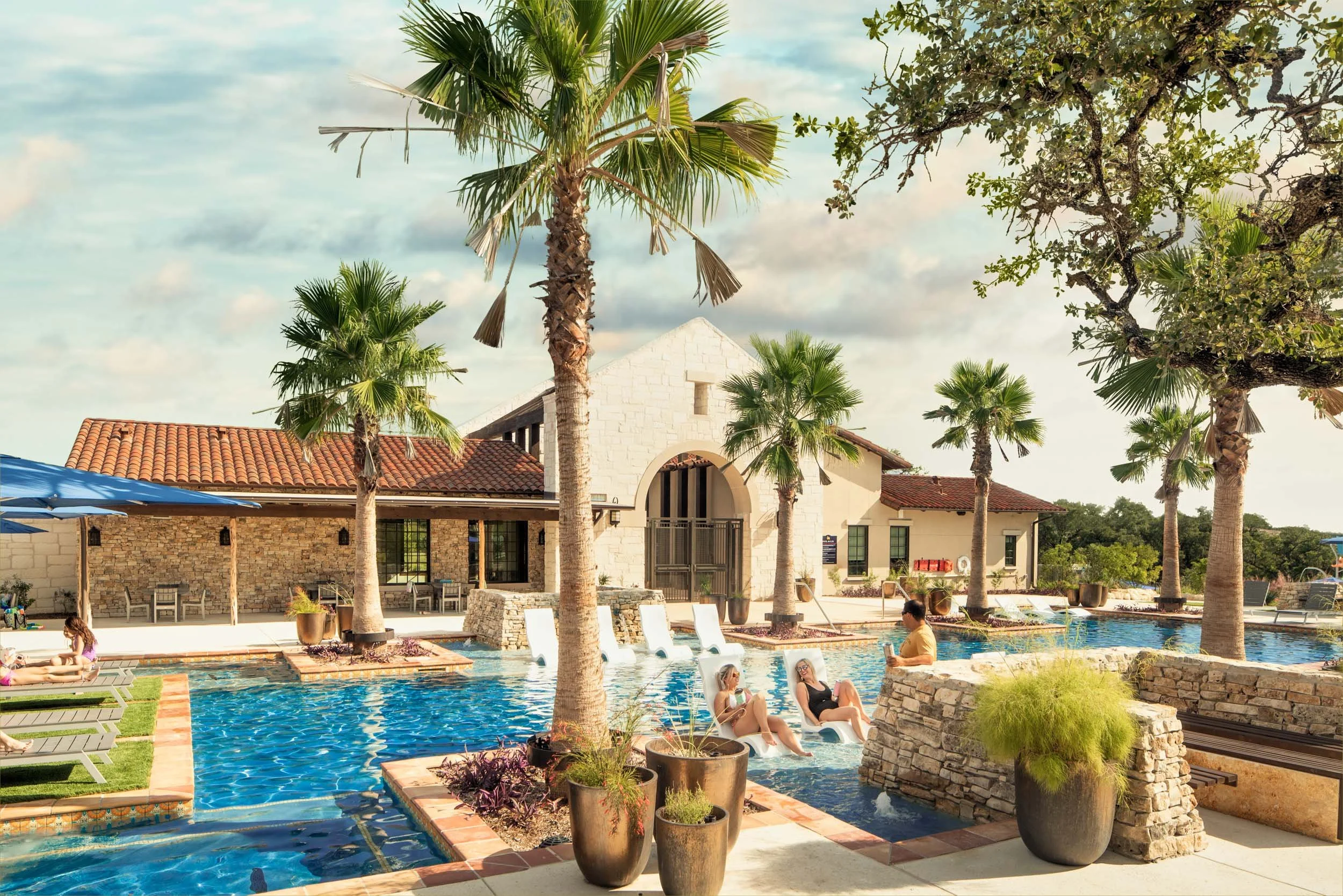 People relaxing on lounge chairs by a swimming pool at a hotel with palm trees and a building with a stone and stucco exterior.
