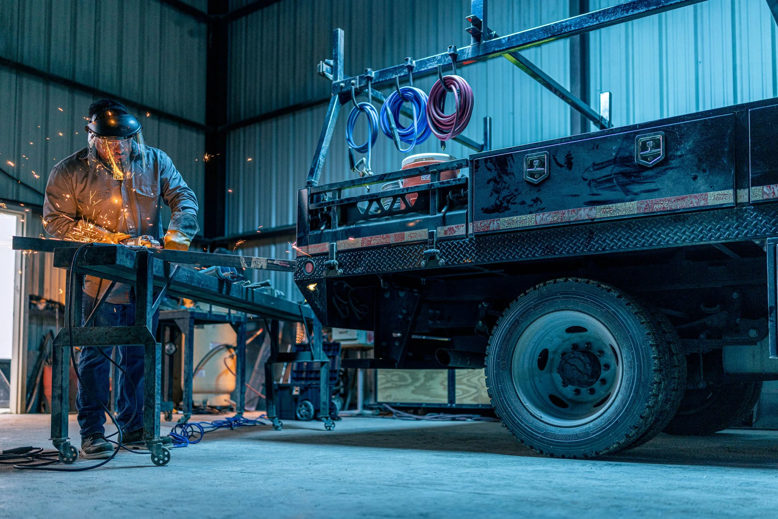 A worker welding inside an industrial garage, sparks flying, near a large black truck with a utility bed and various coiled cables hanging from a rack.