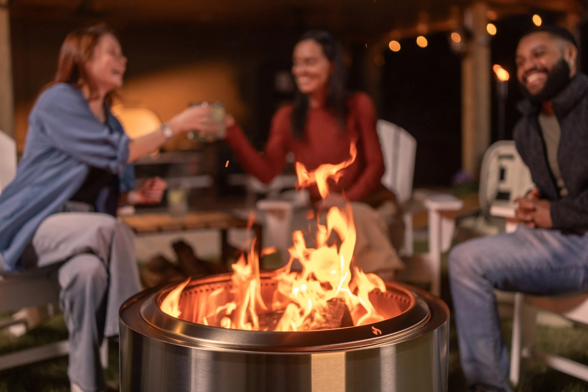 Three people are sitting around a fire pit outdoors at night, laughing and enjoying drinks, with string lights overhead.