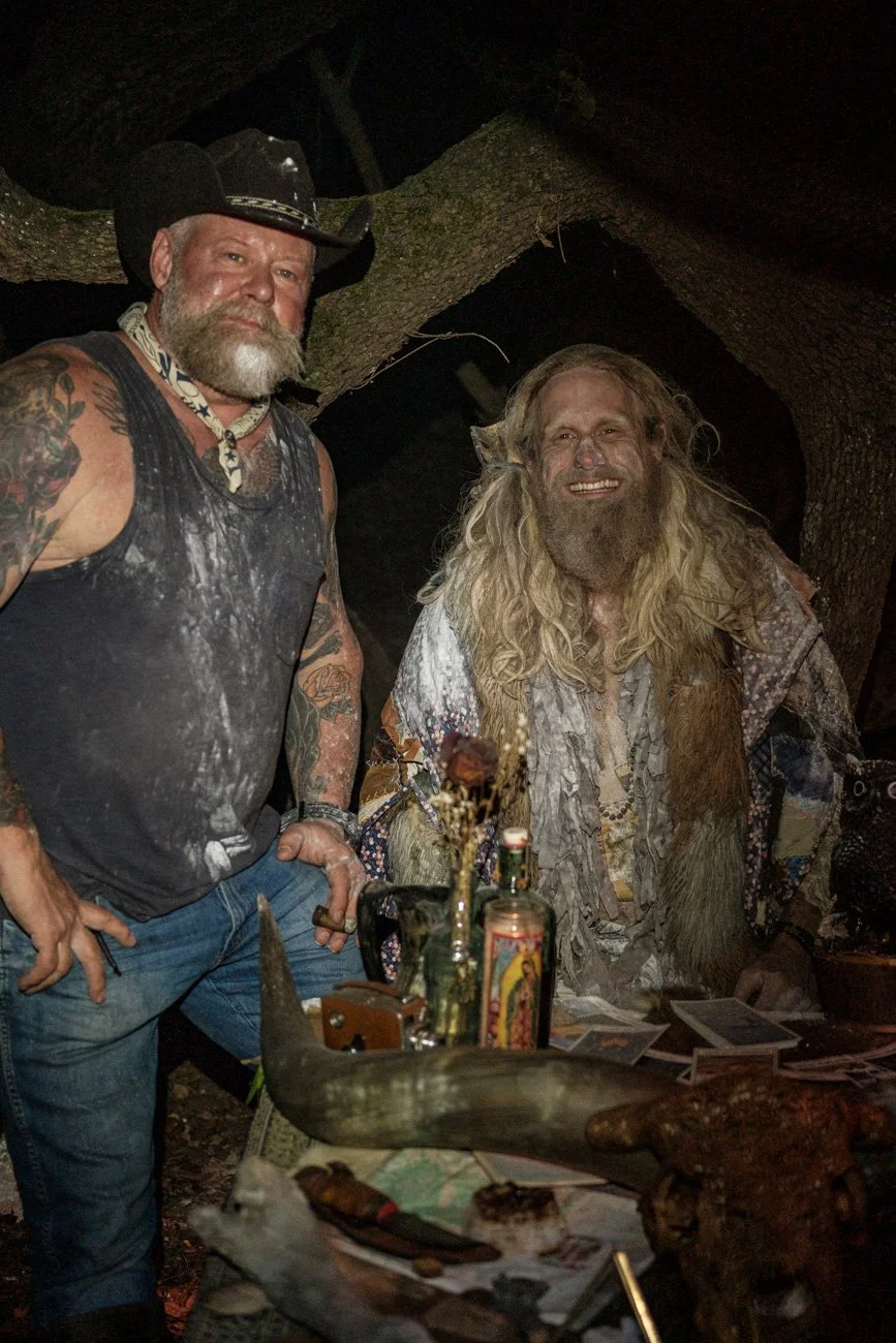 Two men with long hair and beards, one wearing a cowboy hat and sleeveless shirt with tattoos, are inside a dark, wooded area. One is standing with his hand on his hip, the other is seated at a table with candles, photos, and various objects, and bot