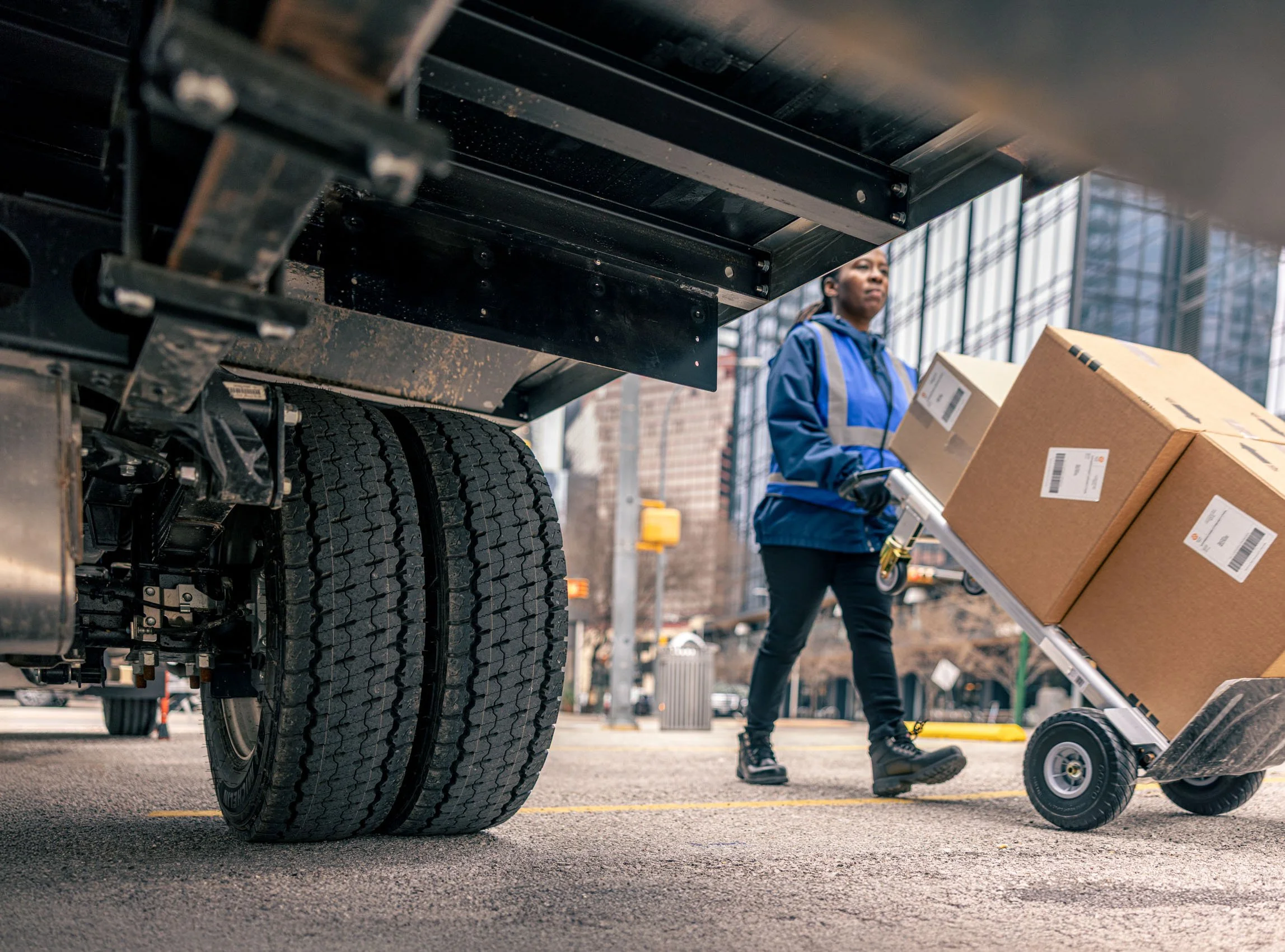 A person in a blue jacket and black pants is moving boxes on a flat hand truck at a city outdoor pickup area. The scene is viewed from underneath a large vehicle with dual tires.