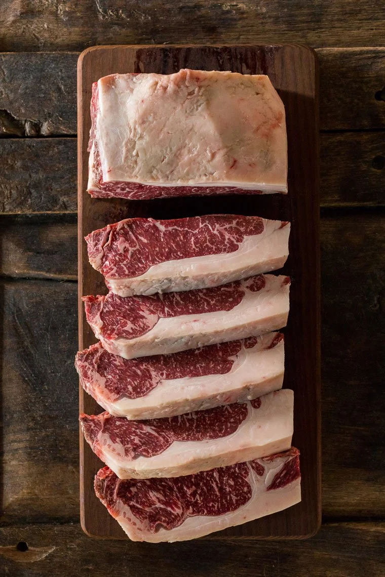 Five cuts of marbled beef steak on a wooden cutting board placed on a dark rustic wooden surface.