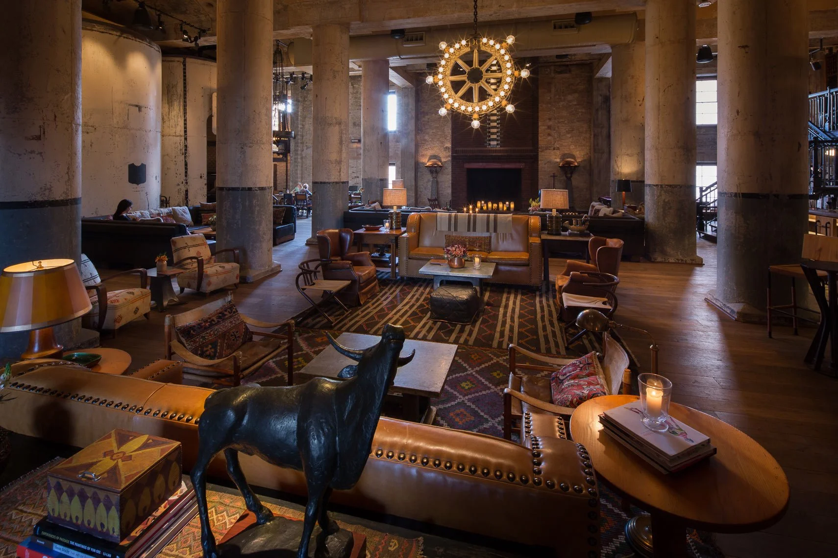Luxurious hotel lobby with high ceilings, large stone columns, and eclectic furniture including sofas, armchairs, and tables. Decor features a large circular chandelier, a fireplace with candles, and various decorative items like books, candles, and 