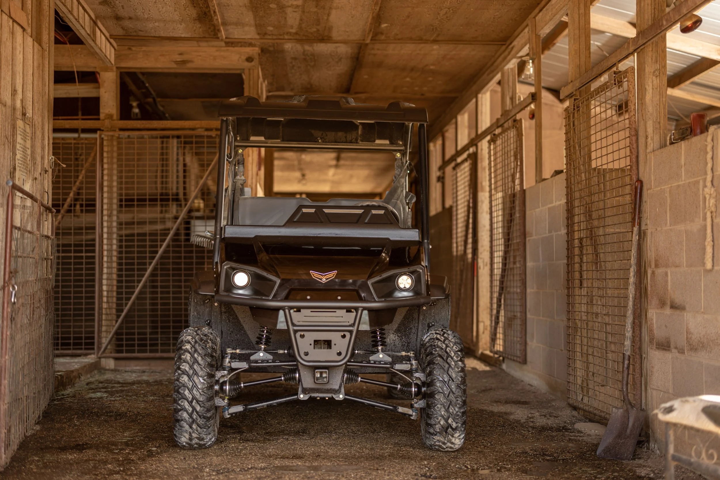 A black off-road utility vehicle inside a barn or garage. The vehicle has large tires, a front grille, and a roof. Surrounding the vehicle are wooden walls, metal gates, and a concrete floor.
