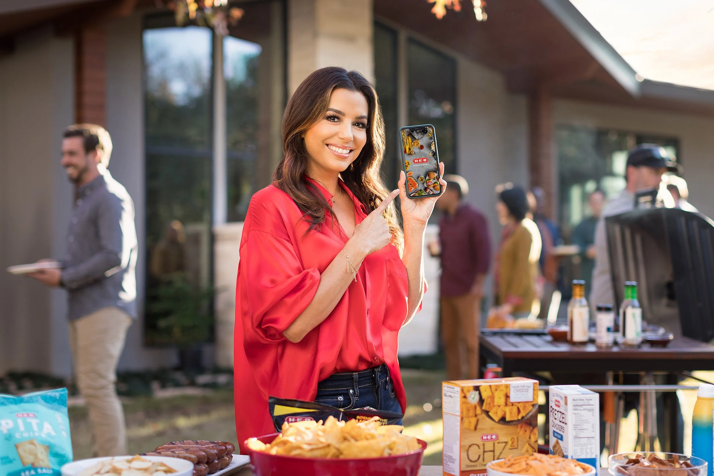 A woman with long brown hair wearing a red blouse is smiling and pointing at her phone, showing a picture of food at an outdoor gathering with several people and a house in the background.