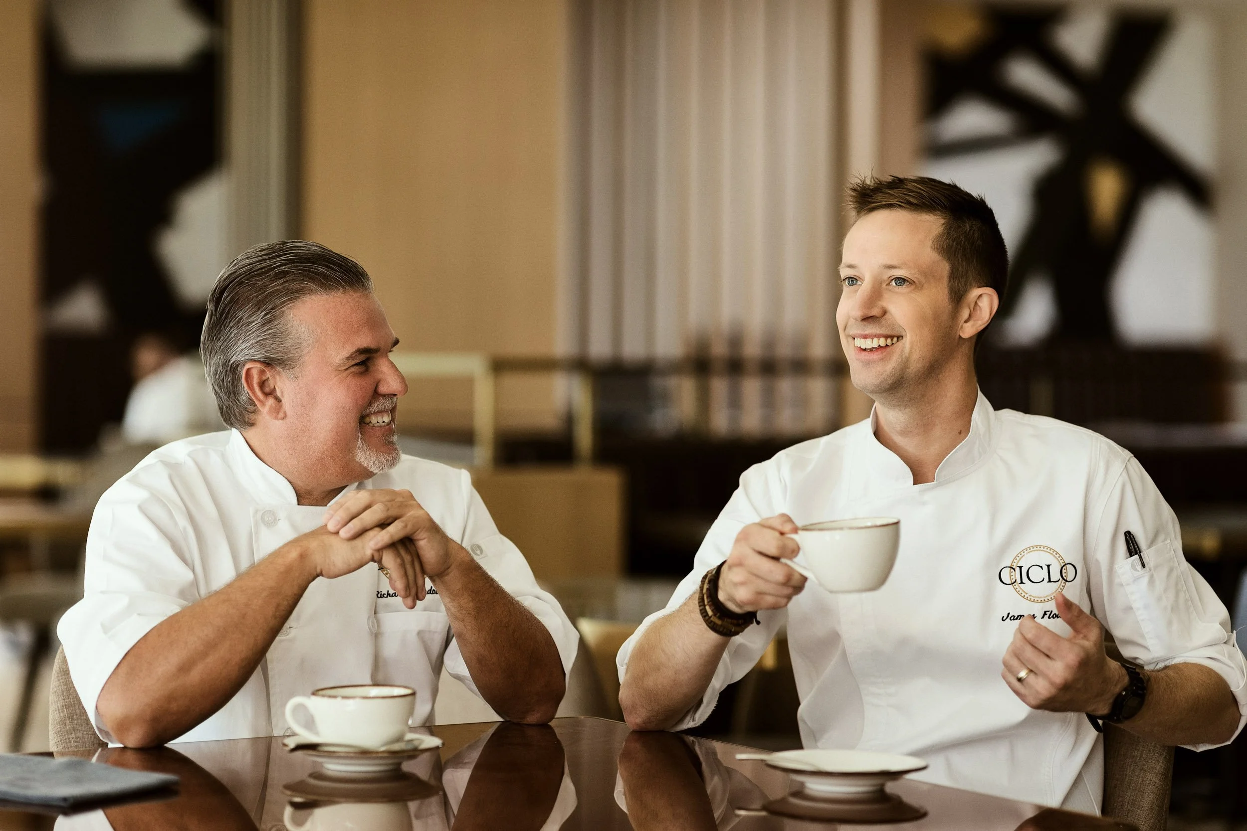 Two male chefs in white uniforms sharing a moment over coffee in a kitchen or dining area, with one holding a white cup and the other resting his hands on the table.