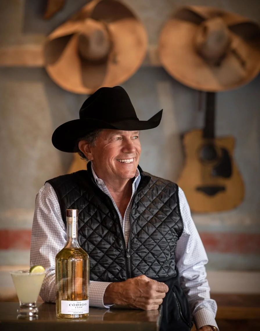 A man in a cowboy hat and quilted vest smiling inside a rustic bar, with a bottle of alcohol and a lemon-lime drink in front of him, and guitars hanging on the wall behind him.