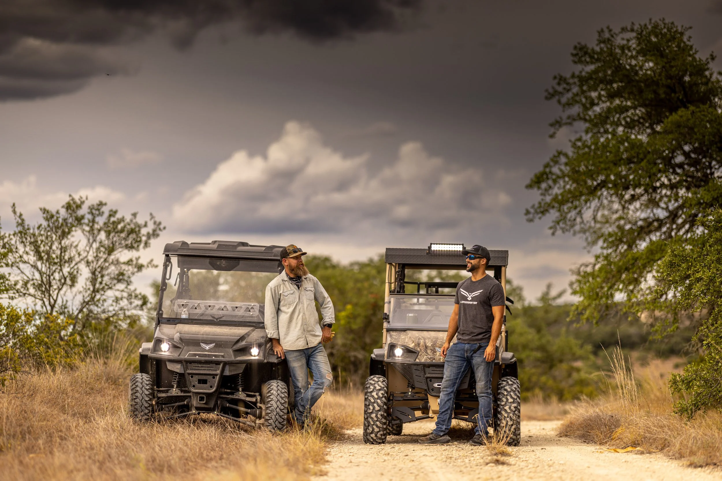 Two men stand and talk on a dirt path, each in a utility vehicle, with dark clouds overhead and trees in the background.