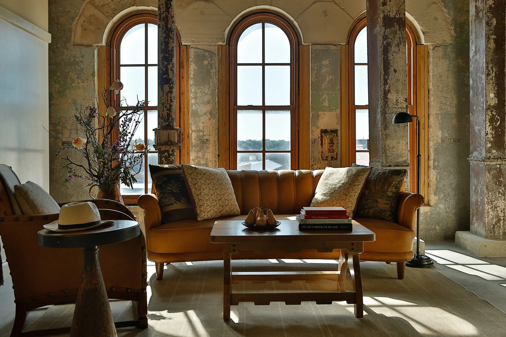 Living room with three large arched windows allowing natural light. Contains a leather sofa with patterned pillows, a wooden coffee table with books and pear-shaped ornaments, an armchair with a hat on it, a metal floor lamp, a flower vase with branc