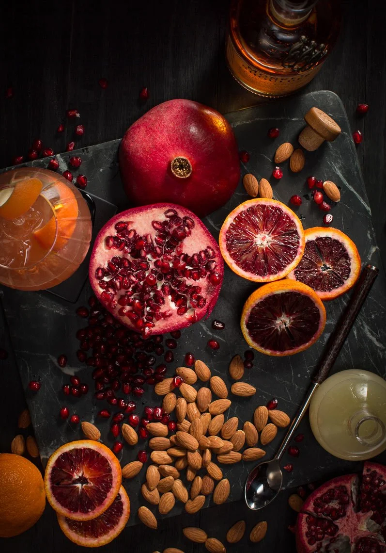 Assorted fruits, including pomegranate, blood oranges, and an orange, with almonds, pomegranate seeds, and a glass of drink on a dark surface.