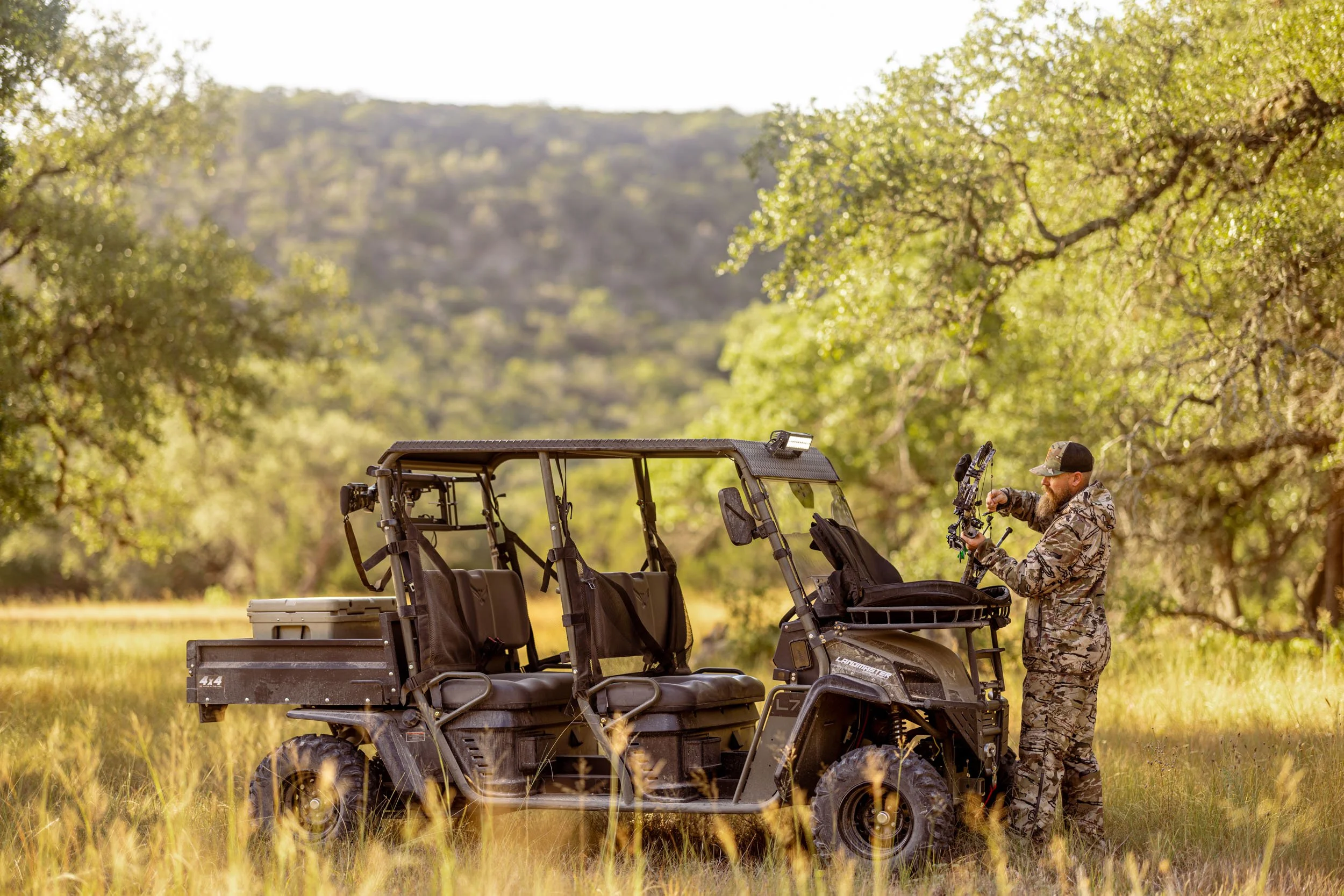 A man in camouflage clothing examining a bow next to a utility vehicle in a field surrounded by trees with green leaves.