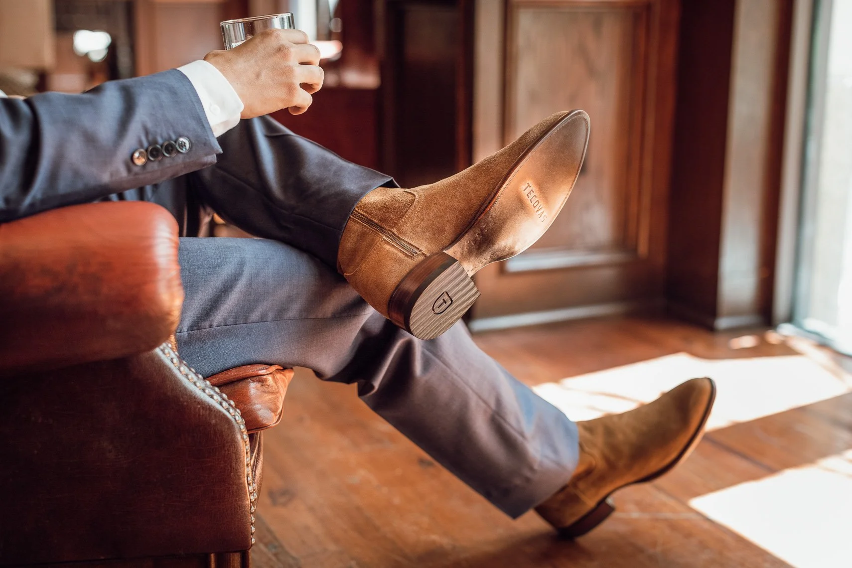 A man sitting comfortably on a leather armchair with his legs crossed, holding a glass of water. He is wearing a suit, and his brown suede boots are prominently displayed, with the sole of one boot showing the brand name TEELONG.
