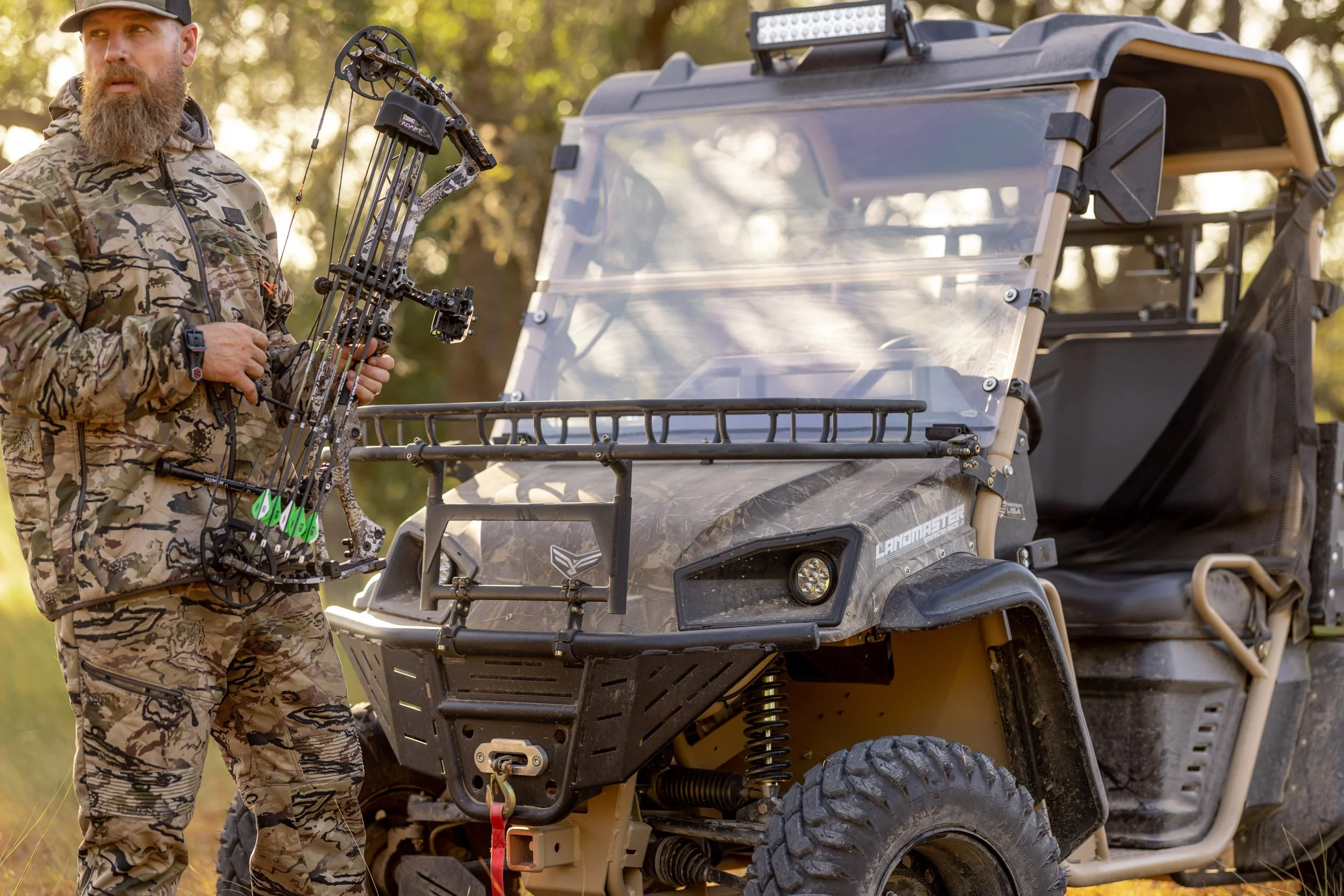 A man dressed in camouflage outdoor clothing holding a compound bow next to an all-terrain vehicle in a wooded area.