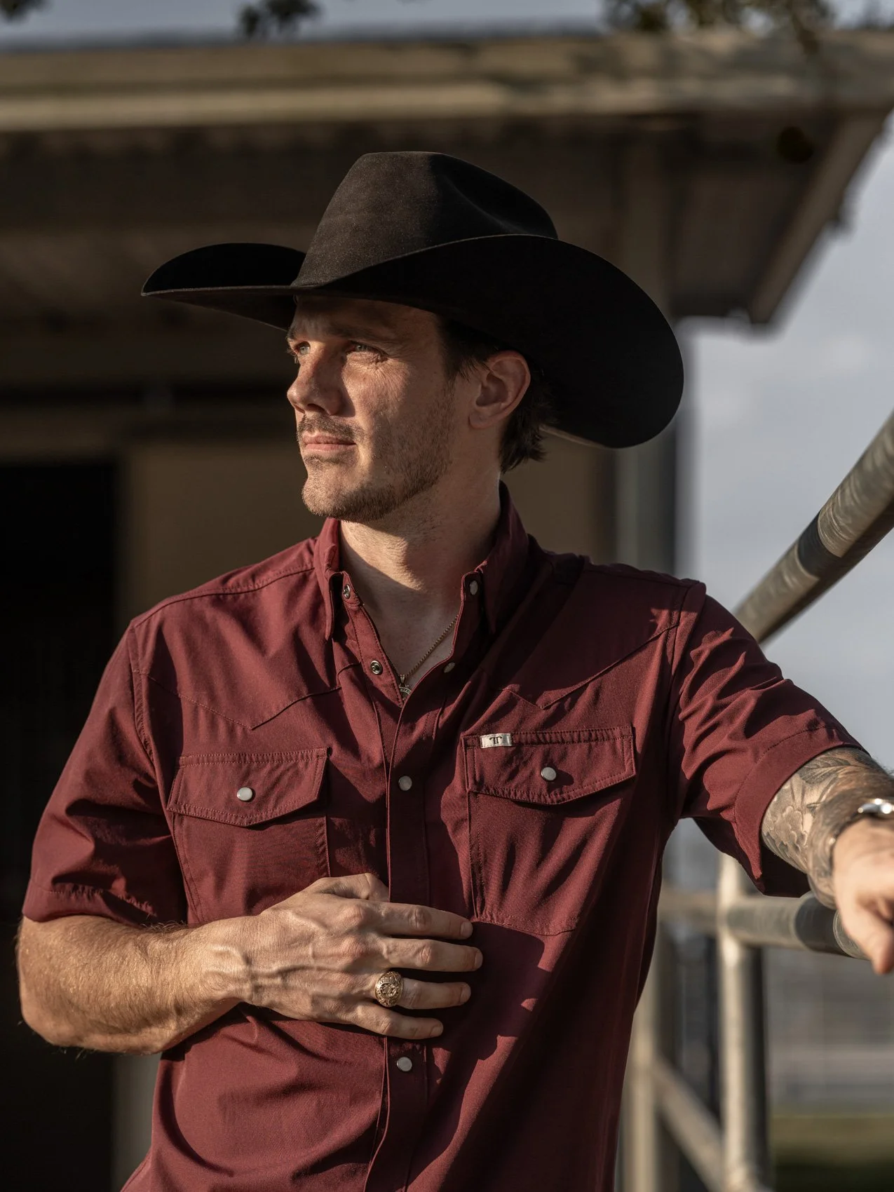 A man in a cowboy hat and red shirt looks into the distance while holding his chest, standing near a metal railing outdoors.