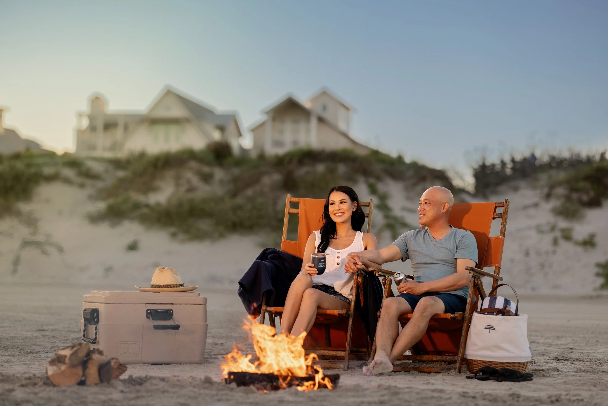 A man and woman sitting on beach chairs near a campfire on the beach, smiling and holding hands, with houses on a hill in the background during sunset.