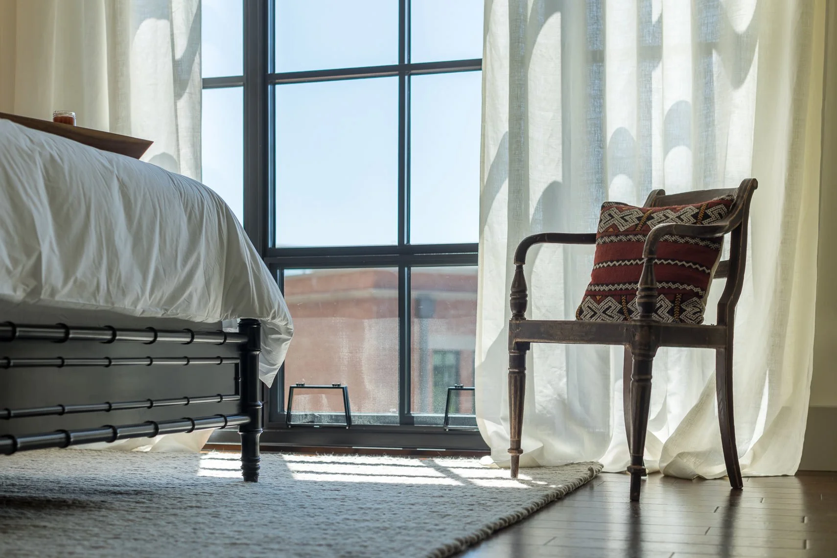A bedroom with a bed on the left, large window with white curtain, and a wooden armchair with a decorative cushion on the right.