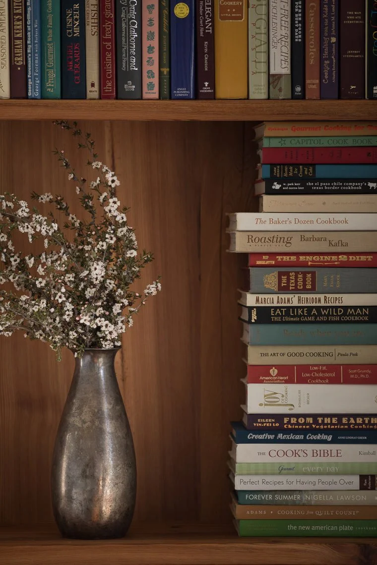 A bookshelf with various cookbooks stacked vertically on the right side and more books on the top shelf. A silver vase containing white and purple flowers is placed on the left side of the shelf.