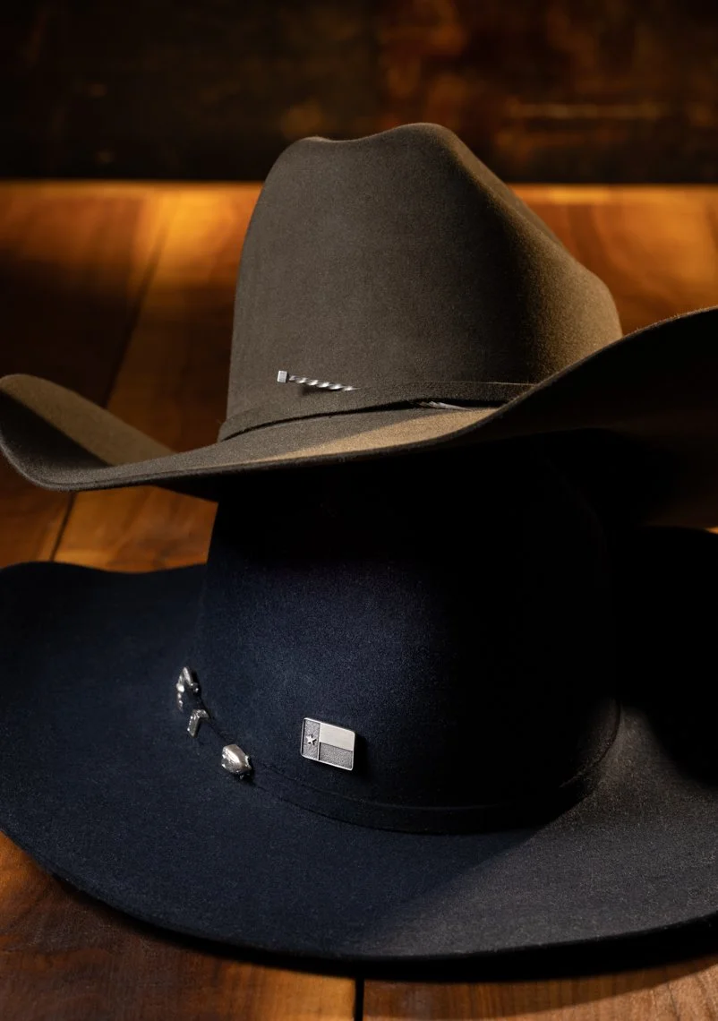 Two cowboy hats, one gray and one black, resting on a wooden surface, with concho and badge accessories attached.