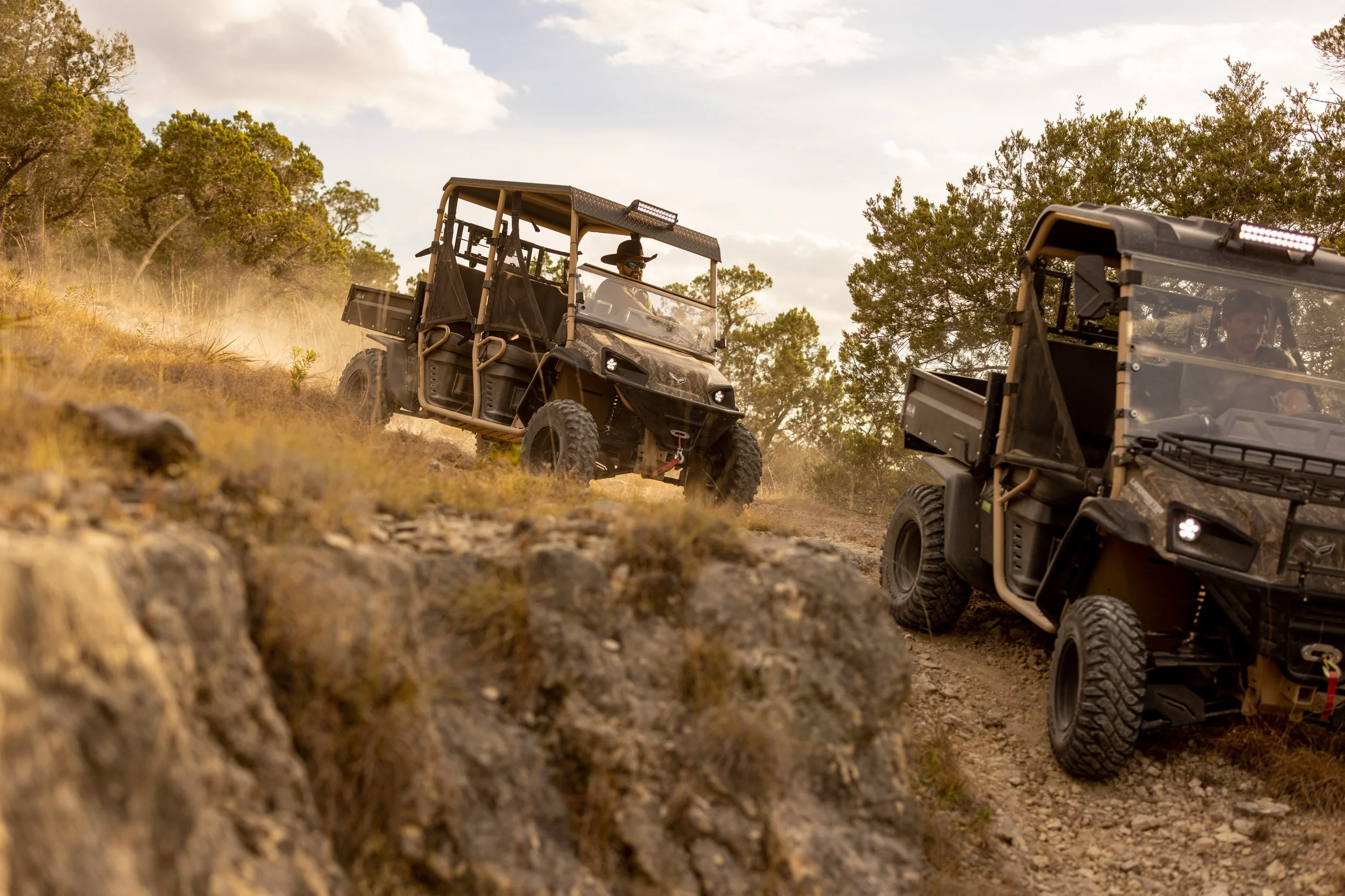 Two off-road utility vehicles driving on a dirt trail in a rural area with trees and a cloudy sky.