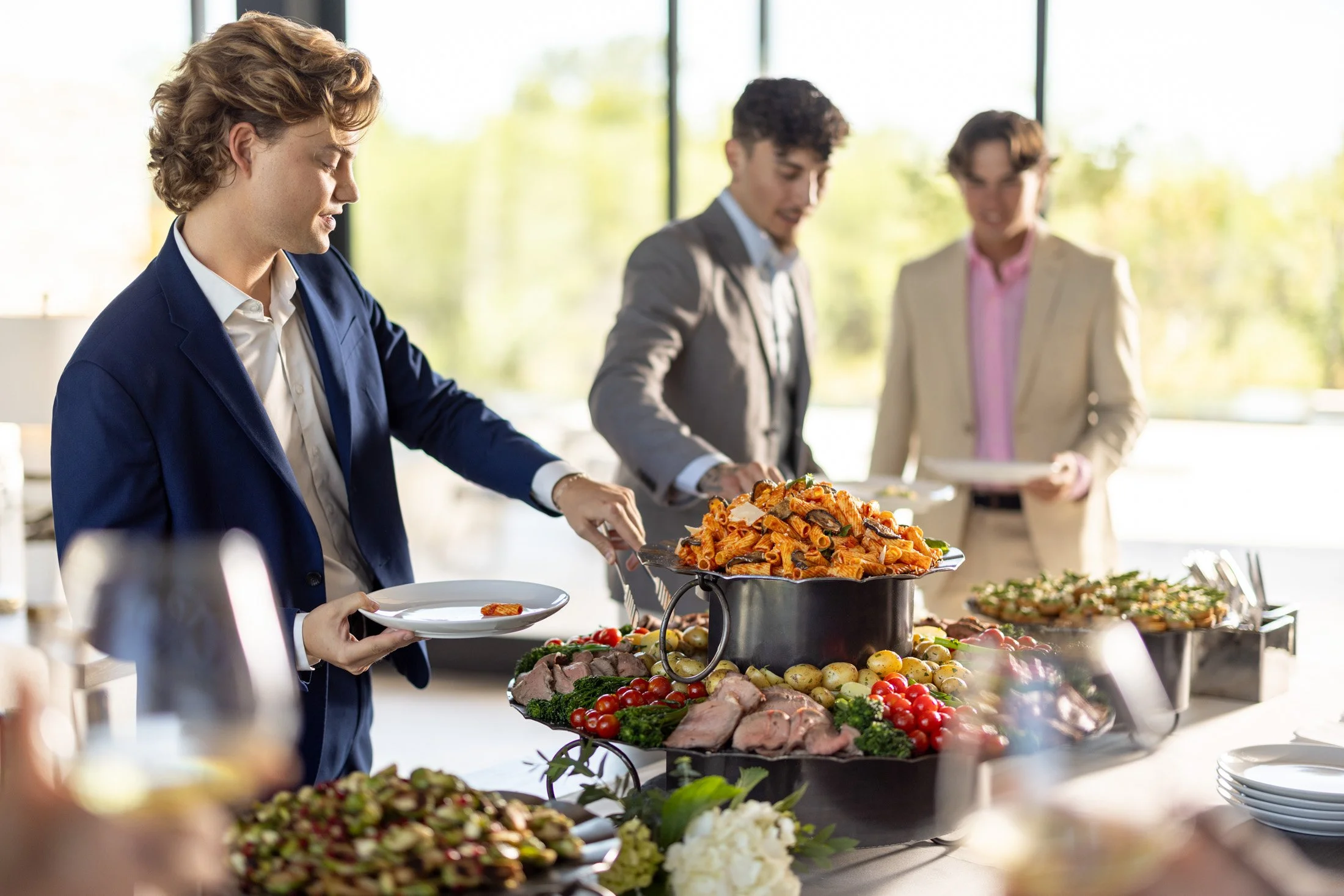 People serving themselves food from a buffet featuring pasta, salads, and vegetables in a bright, modern setting with large windows.