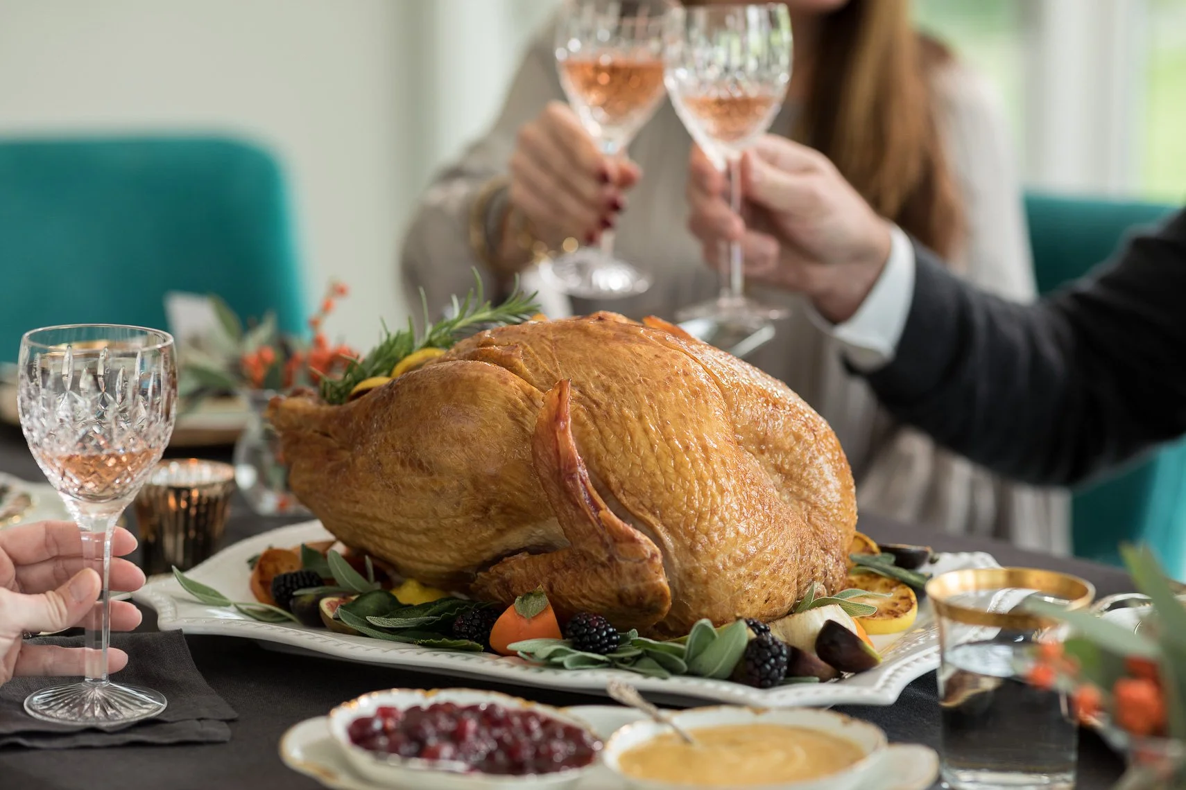 A festive meal with a roasted turkey as the centerpiece, surrounded by garnishes and side dishes, with people raising glasses of rosé wine for a toast.