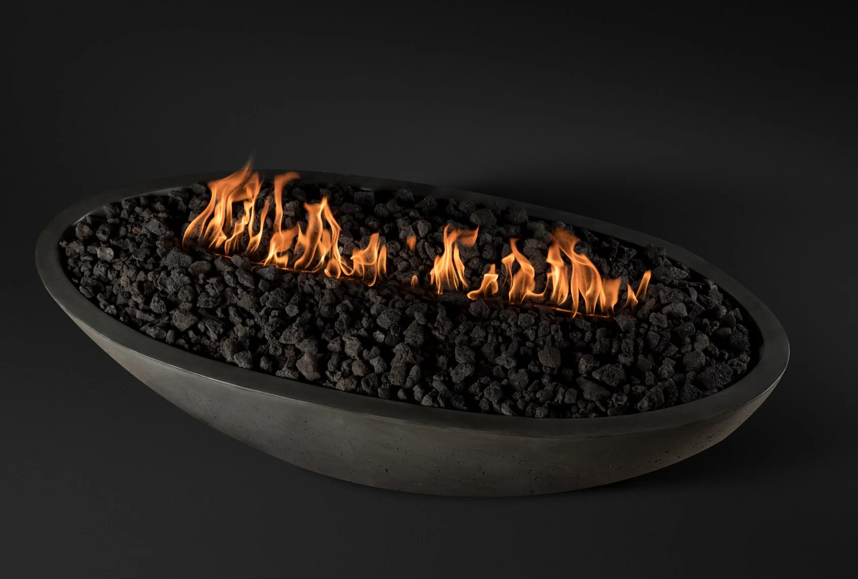 Modern oval fire pit filled with black rocks and lit with small, bright flames against a dark background.