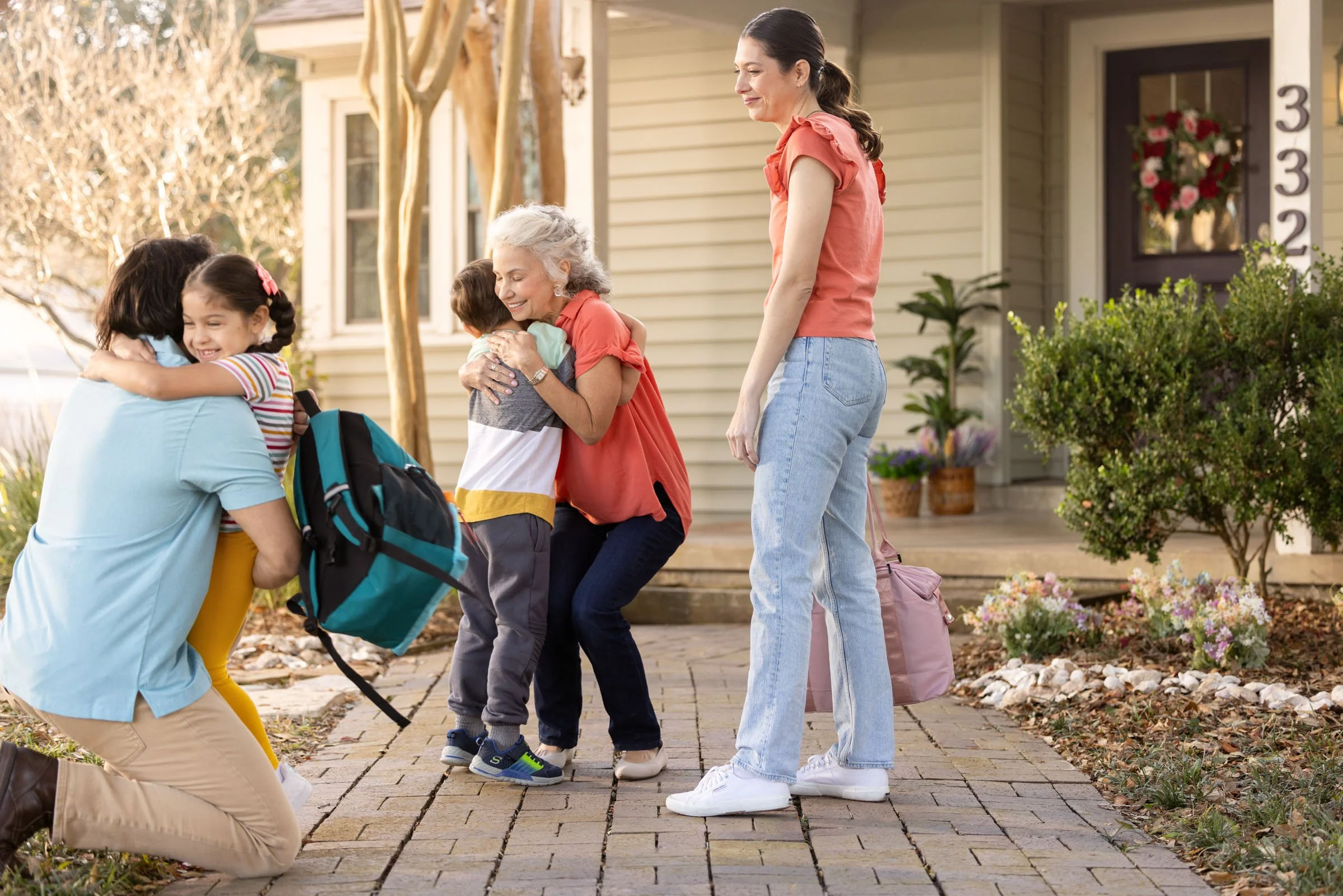 A family is greeting each other with hugs outside a house. An elderly woman, a young woman, and three children are visible. The family appears happy, and the scene is set in a residential neighborhood with plants and flowers around.