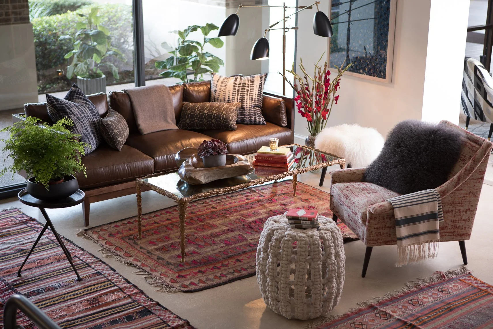 Living room with brown leather sofa decorated with patterned pillows, a glass coffee table with decorative items, a floral upholstered armchair with a pillow, colorful rugs on the floor, indoor plants near large window, and a wall art piece.