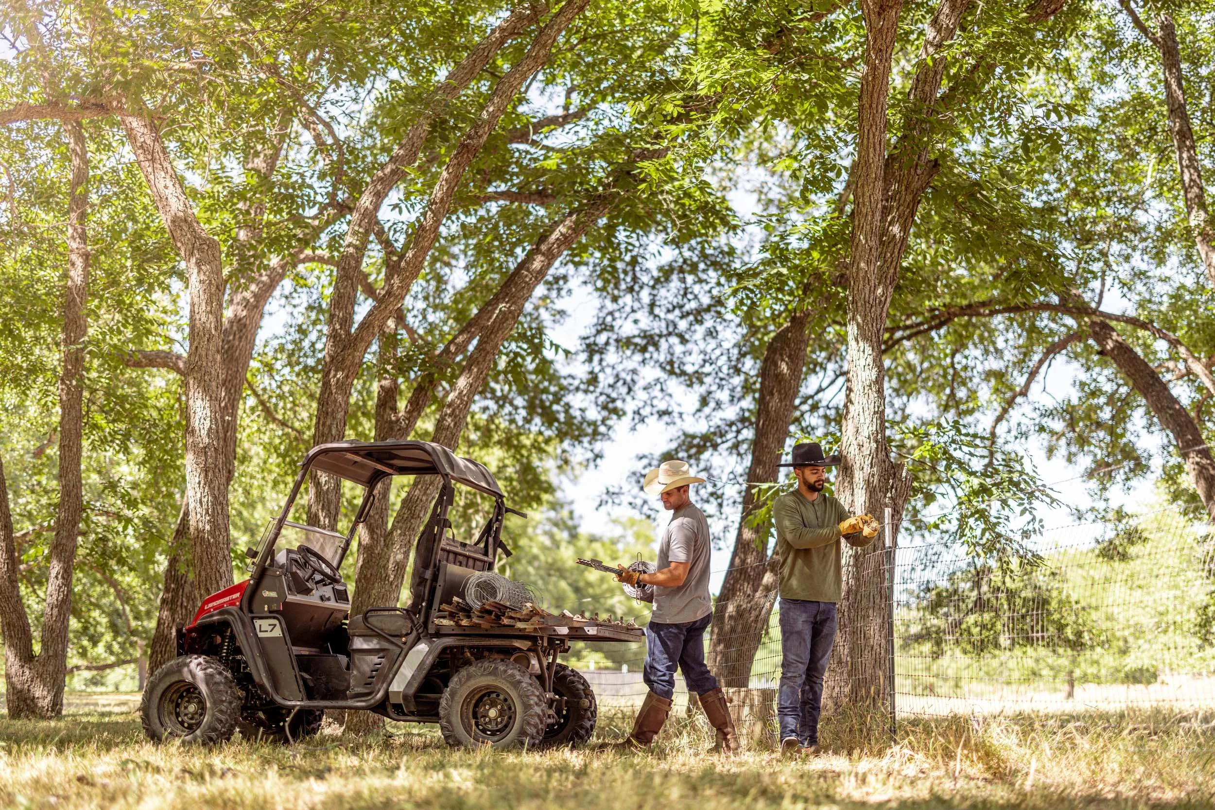 Two men working outdoors among tall trees with green leaves, one in a gray shirt and jeans, the other in a green shirt, both wearing hats, near a utility vehicle and tools.